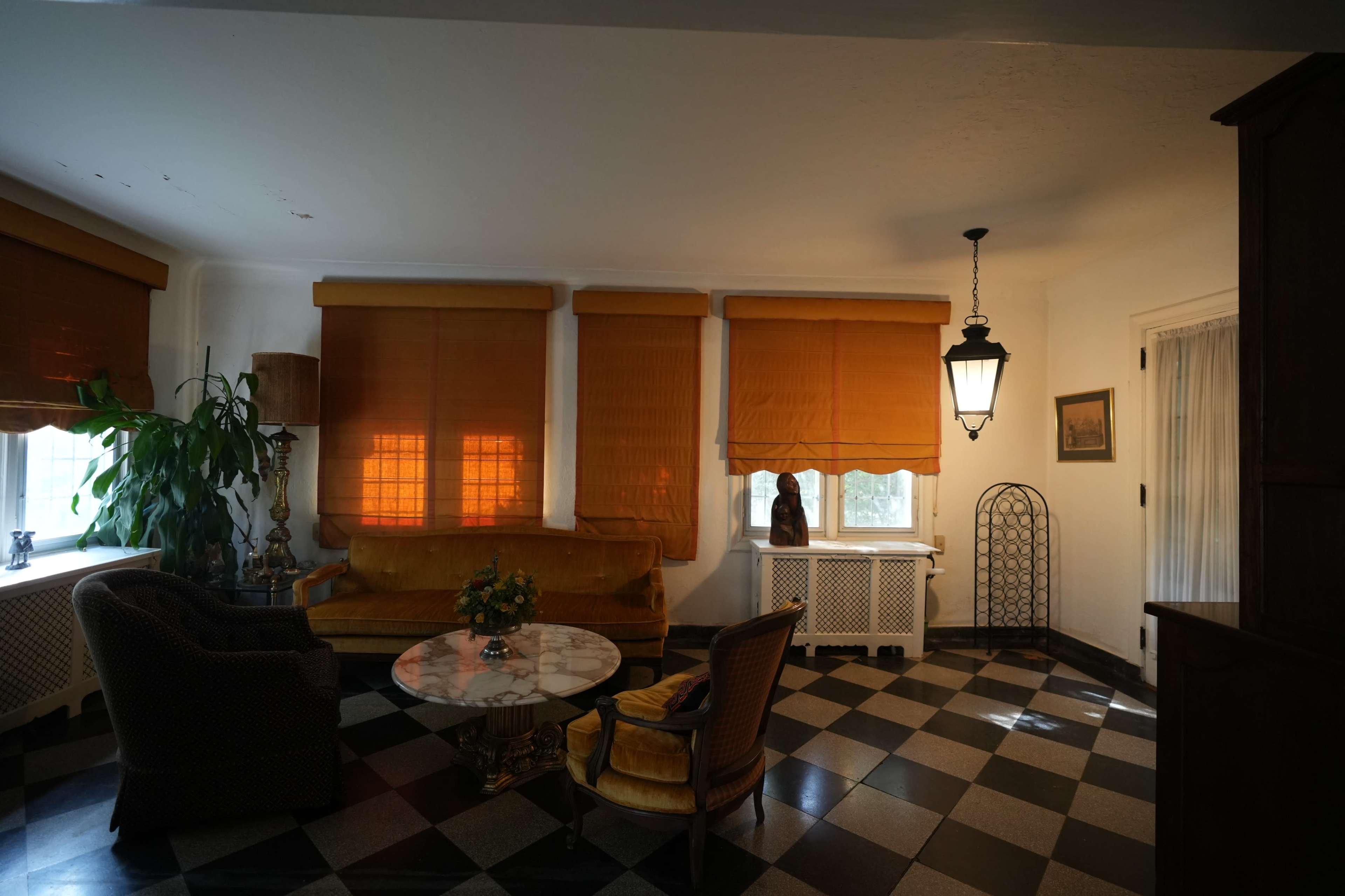 The image shows a room with checkered flooring, a yellow sofa, a small round table with a floral arrangement, and large windows covered with orange curtains.
