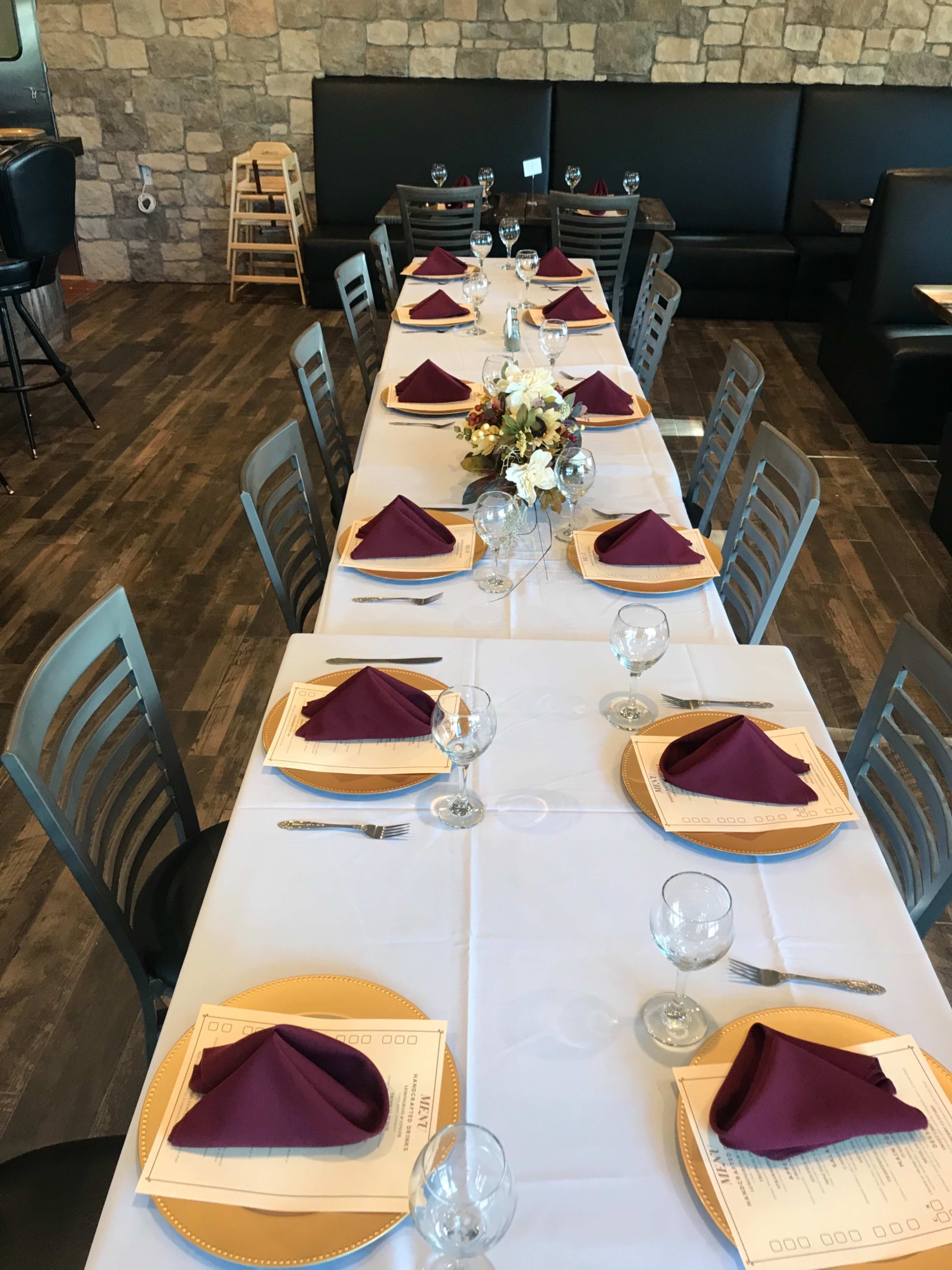 A long dining table is elegantly set with purple napkins, clear glassware, and a floral centerpiece against a backdrop of stone walls and dark seating.