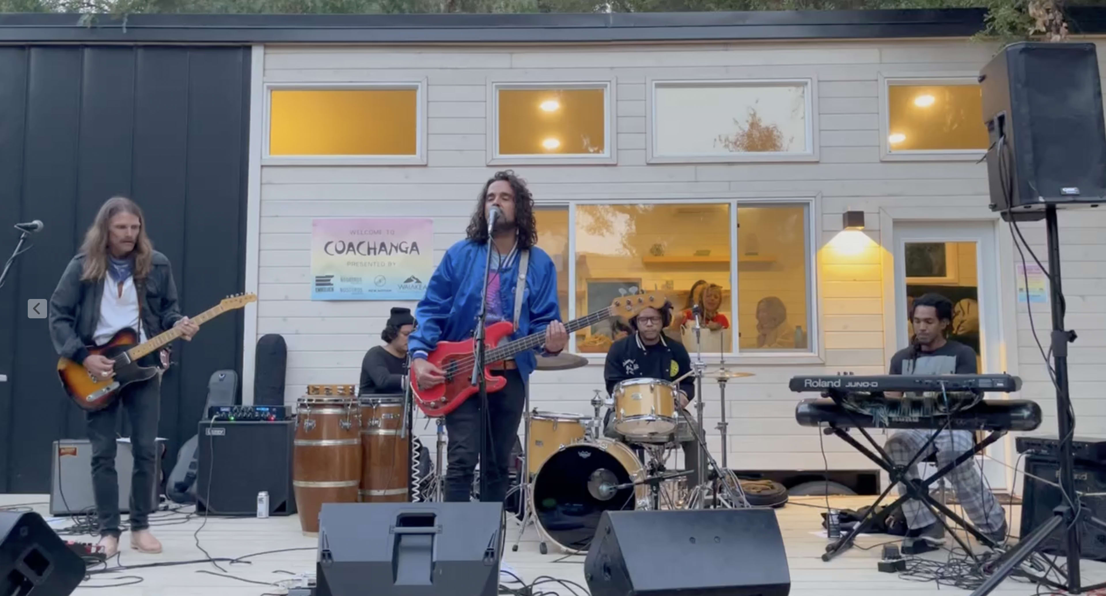 A band performs on an outdoor stage with various instruments, including electric guitars, drums, and a keyboard.