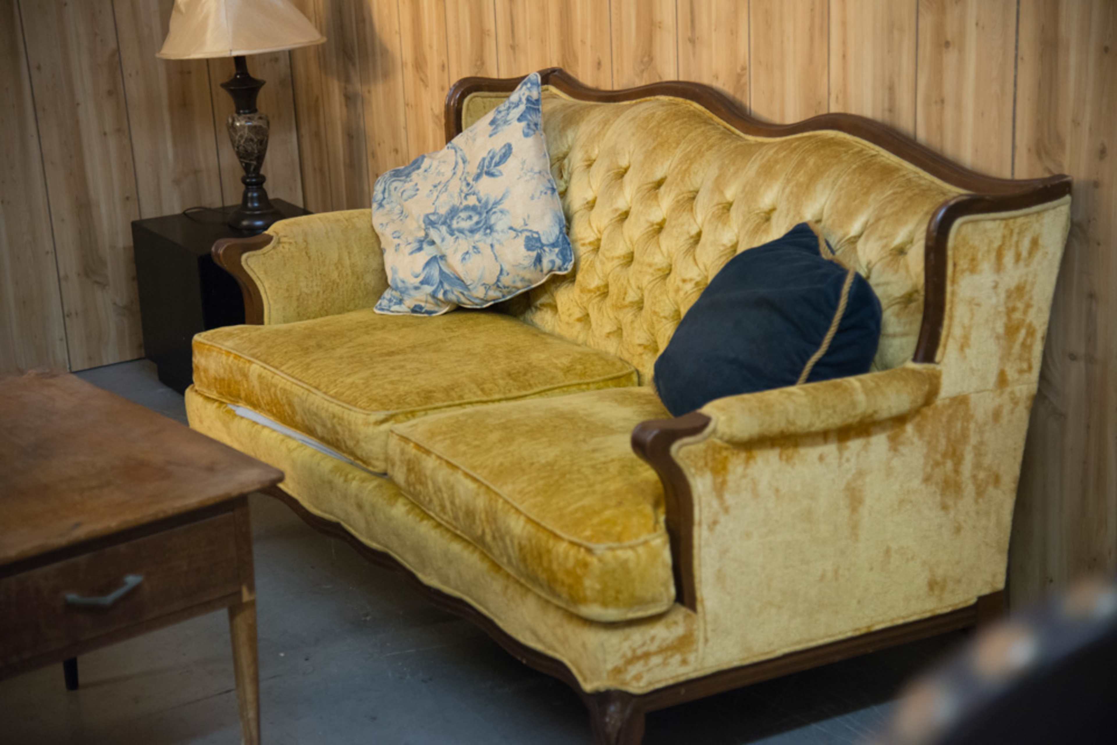 A vintage yellow upholstered sofa with a tufted backrest and two cushions, accompanied by a wooden side table and a lamp in a room with wooden paneling.