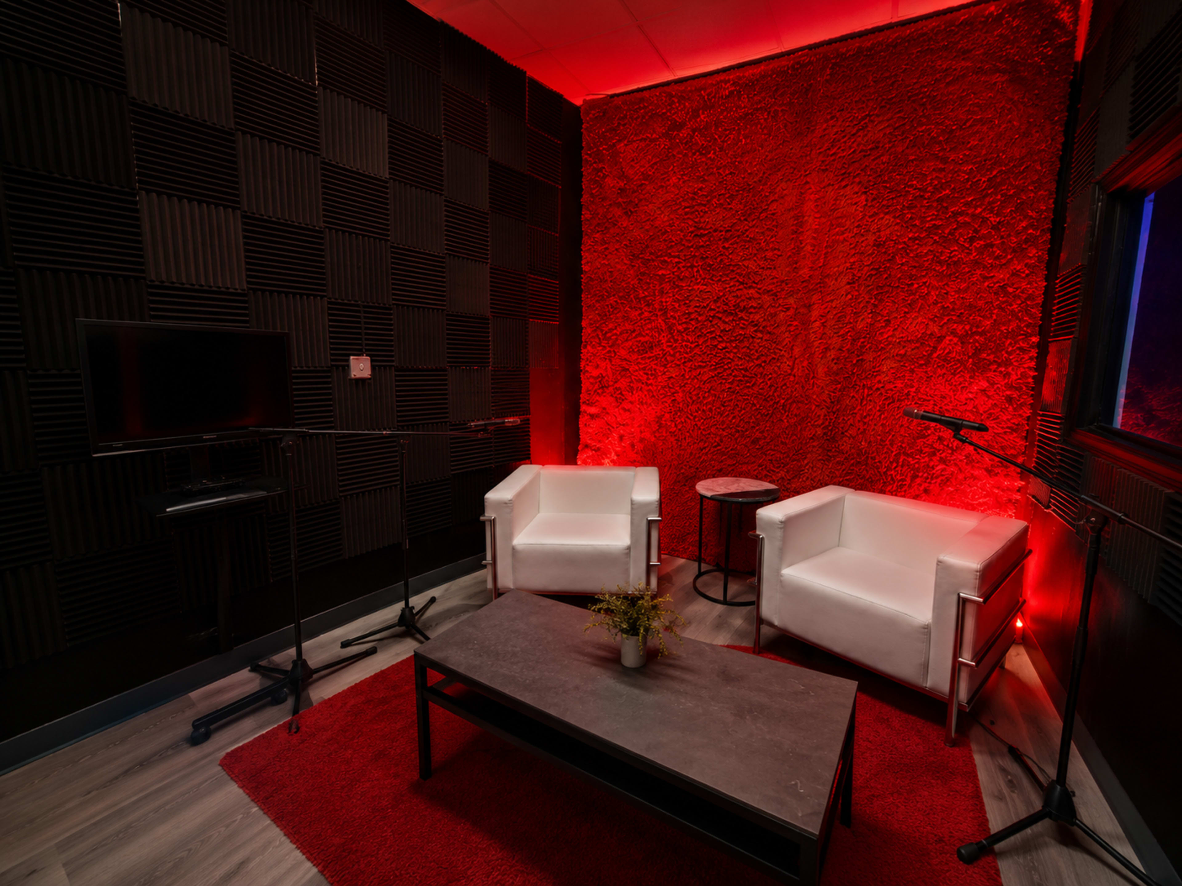 The image shows a small, modern room with black textured walls, red lighting, two white armchairs, a coffee table, and a television on a stand.