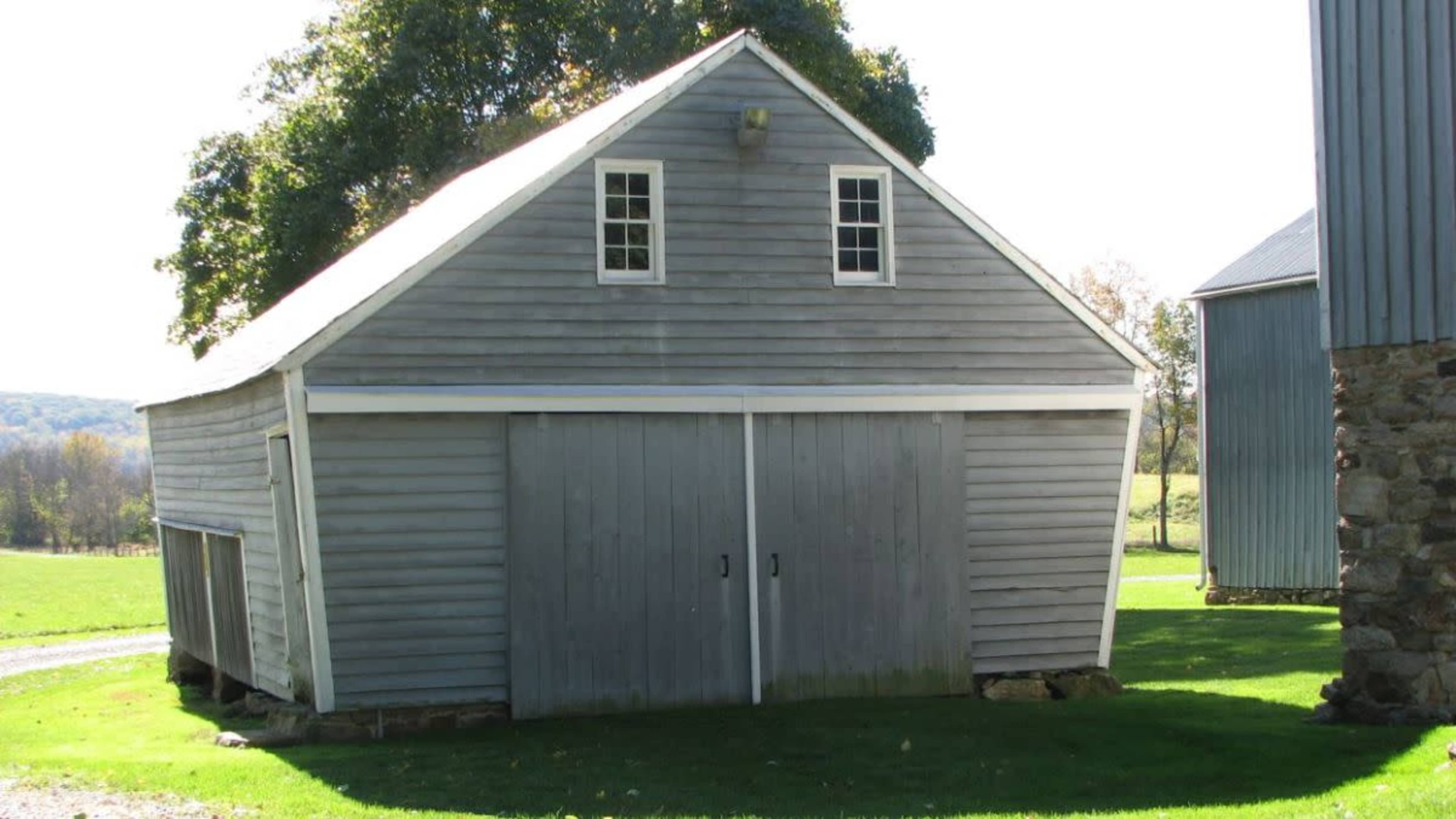 A gray wooden barn with two doors, set on a grassy area beside a gravel path.