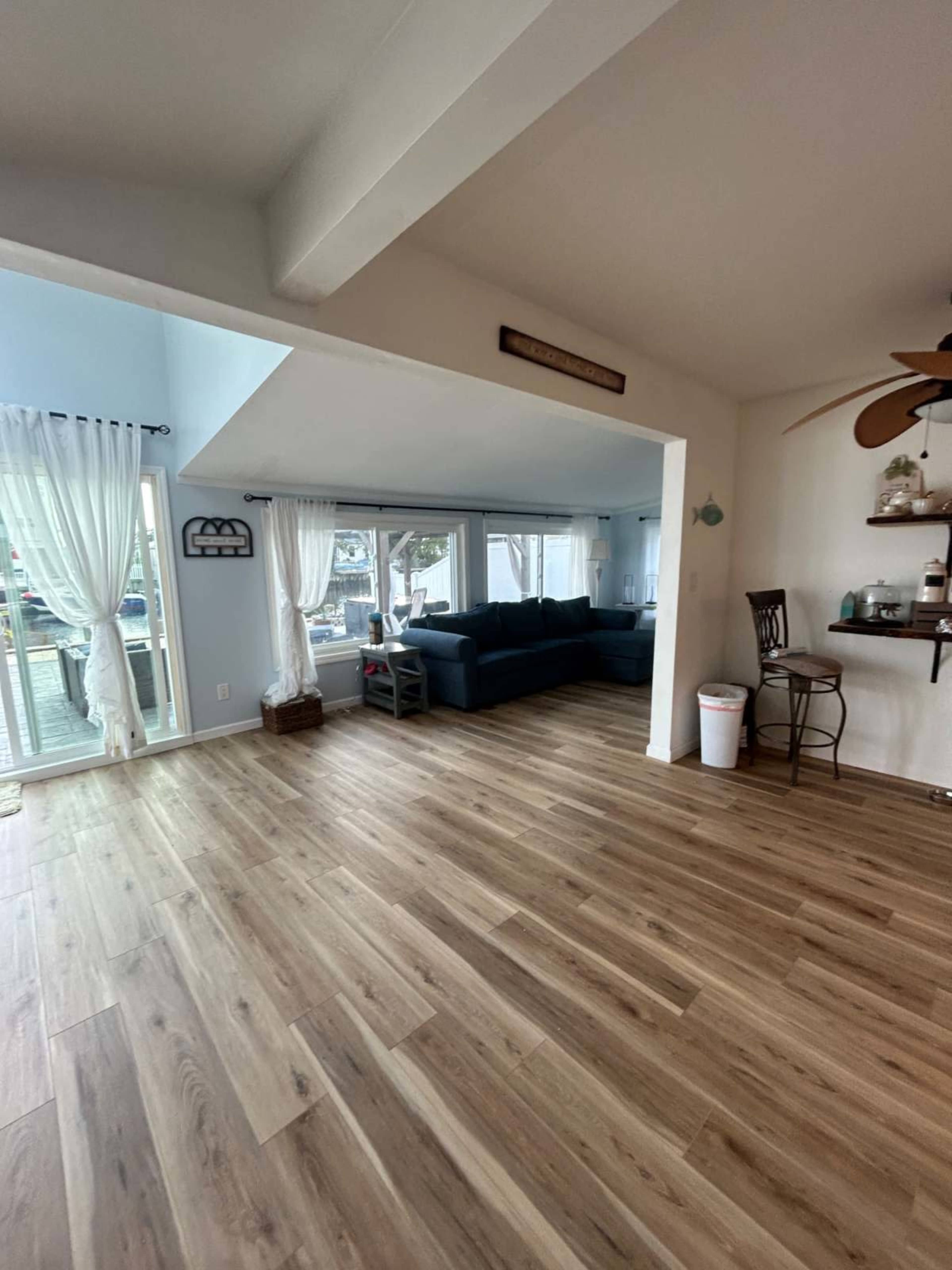 A spacious room features wooden flooring, large windows with sheer curtains, and a seating area with a dark-colored couch.