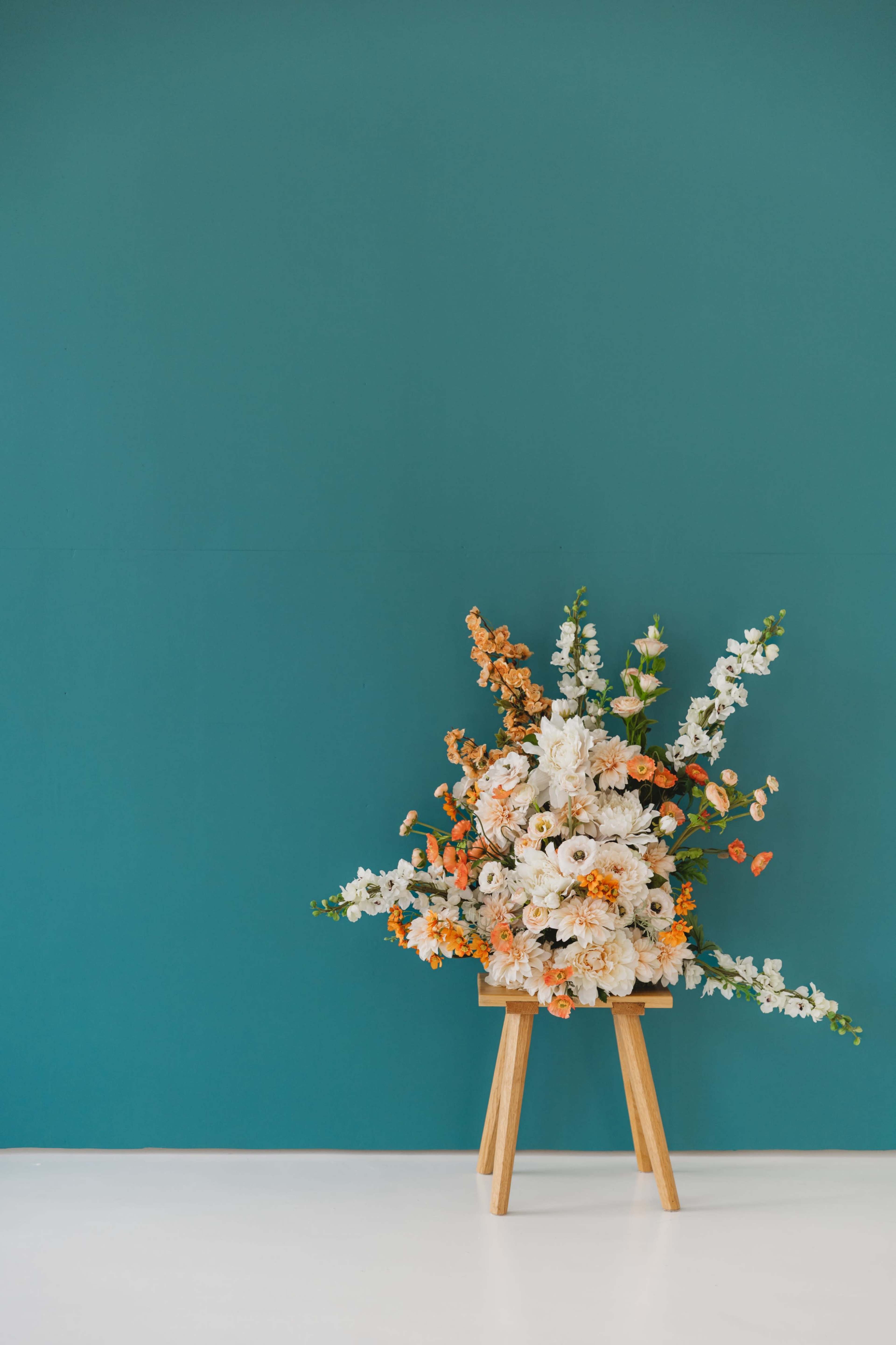 A wooden stand displays a large bouquet of white, orange, and green flowers in front of a teal wall.