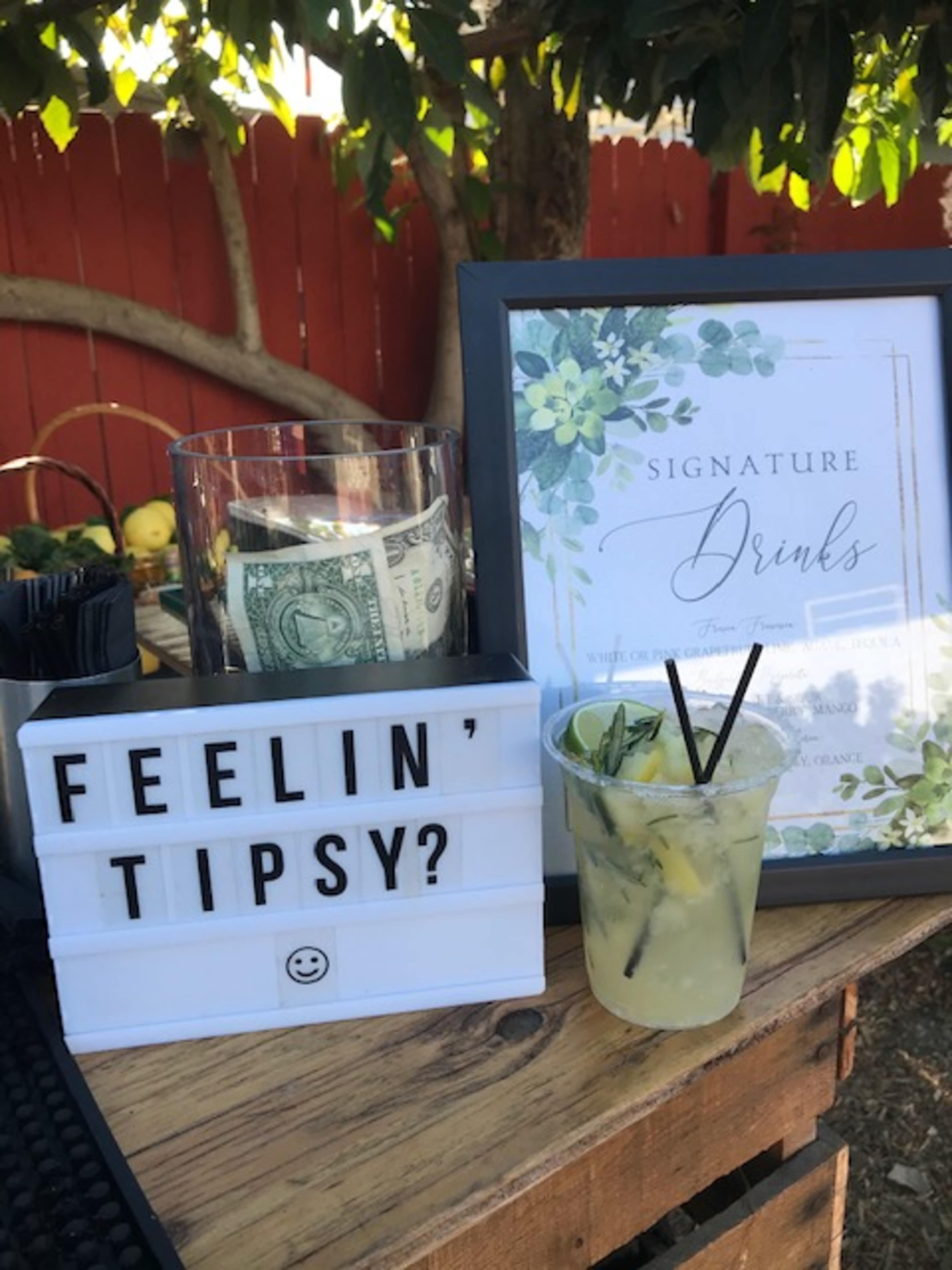 A cocktail sits on a wooden table next to a sign displaying "SIGNATURE DRINKS" and a jar containing a dollar bill.
