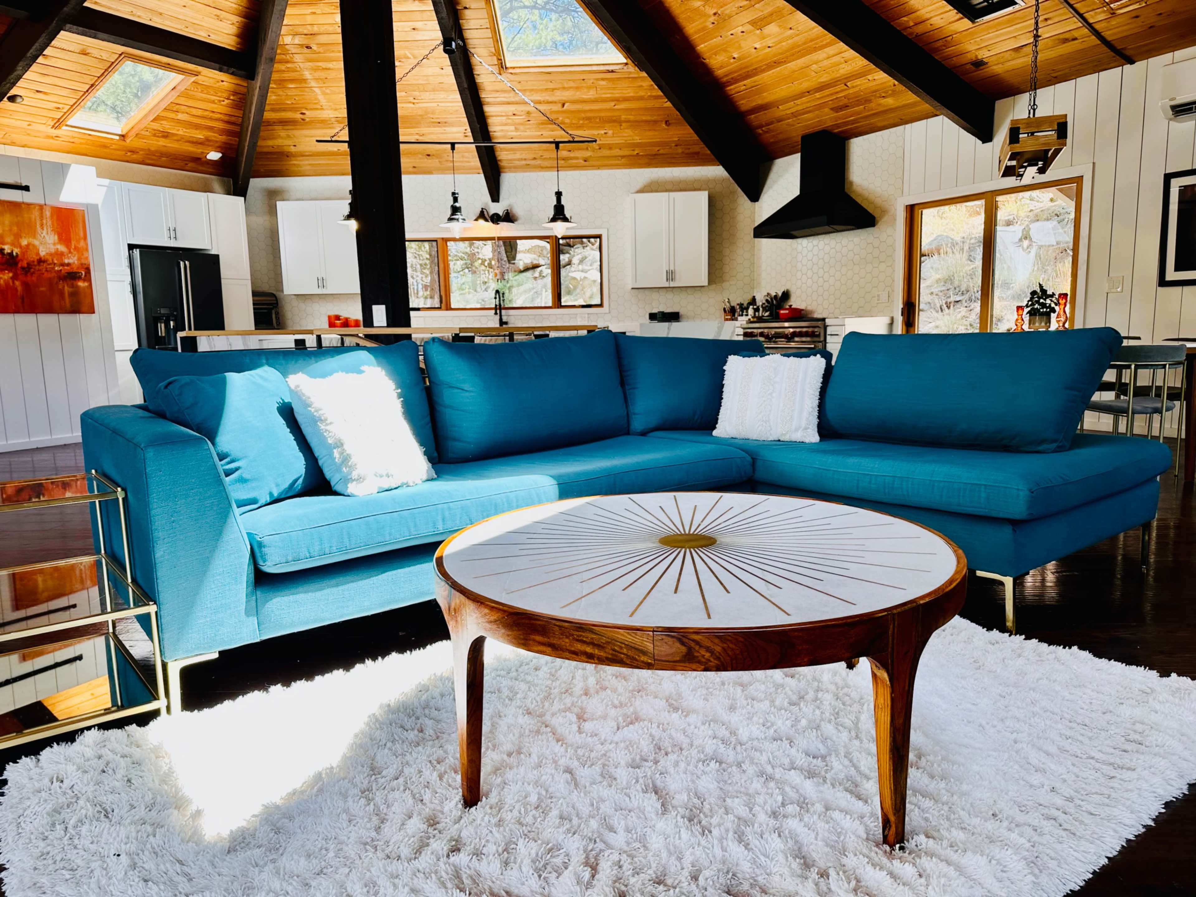 A modern living room with a turquoise sectional sofa, a round coffee table with a sunburst design, and large windows letting in natural light.