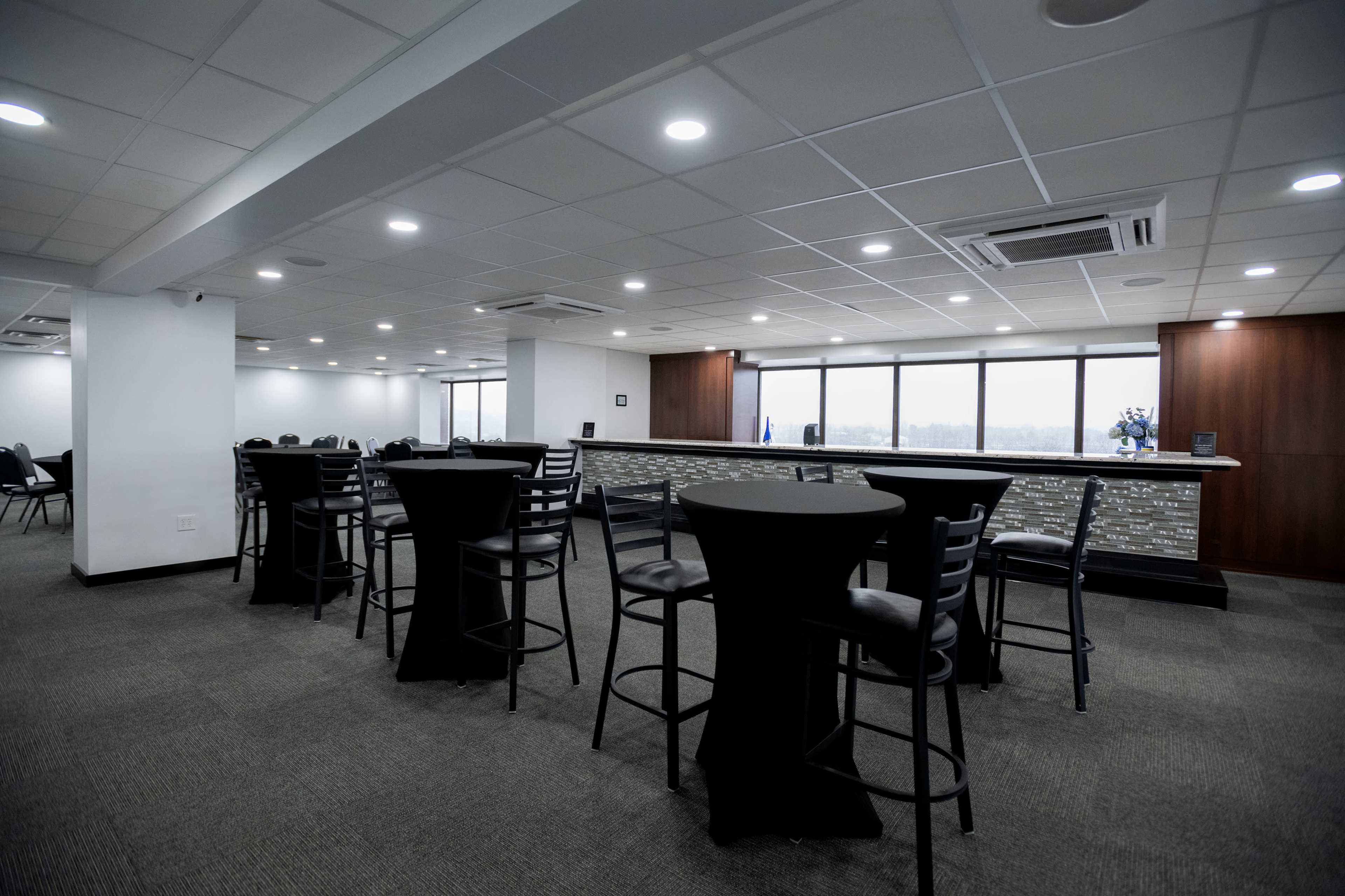 A modern office lounge area with black high-top tables and chairs, well-lit by ceiling lights and natural light from large windows.