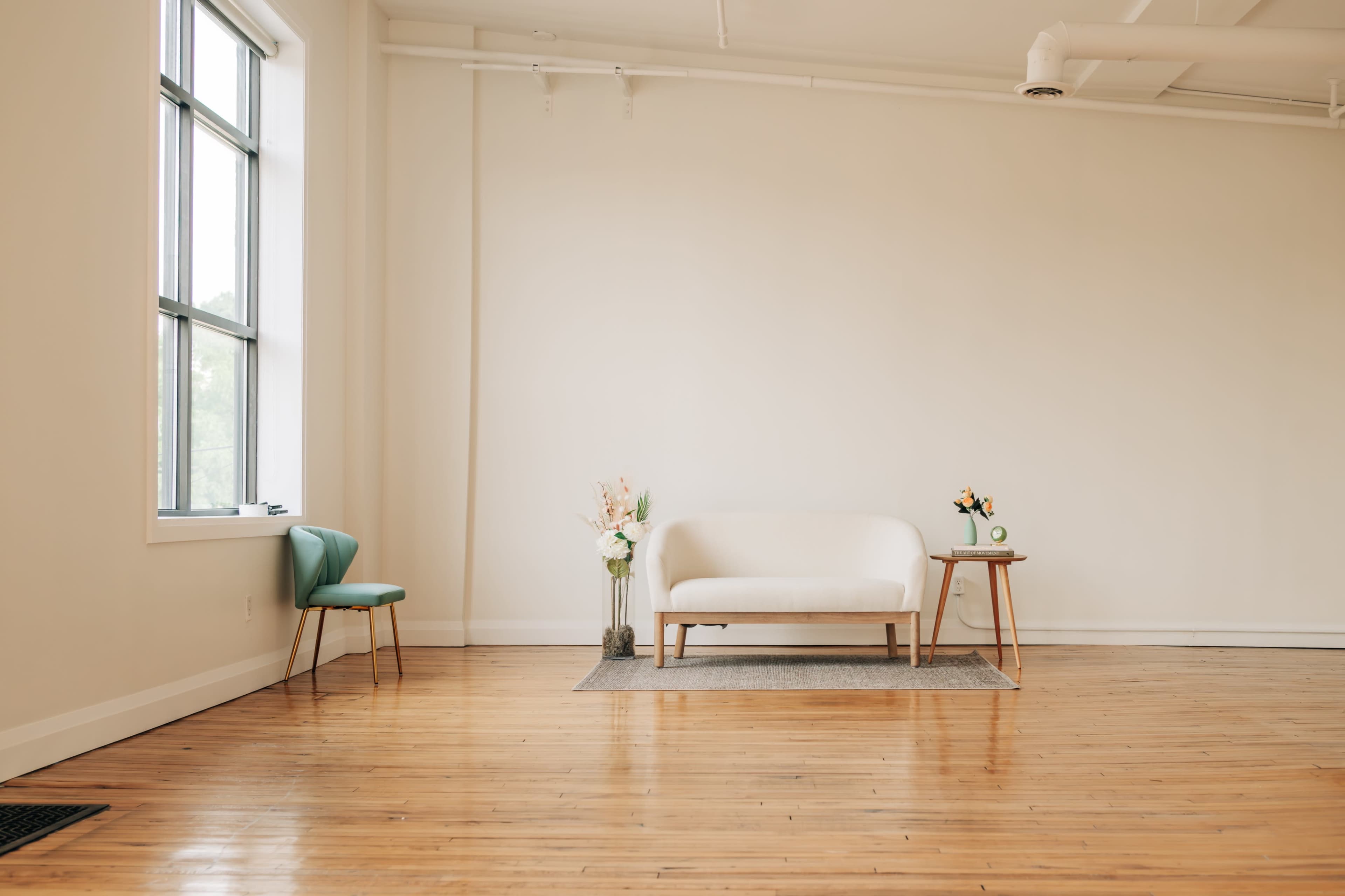 A minimalist room features a white sofa, a small wooden table with a flower vase, an aqua chair, and hardwood flooring.