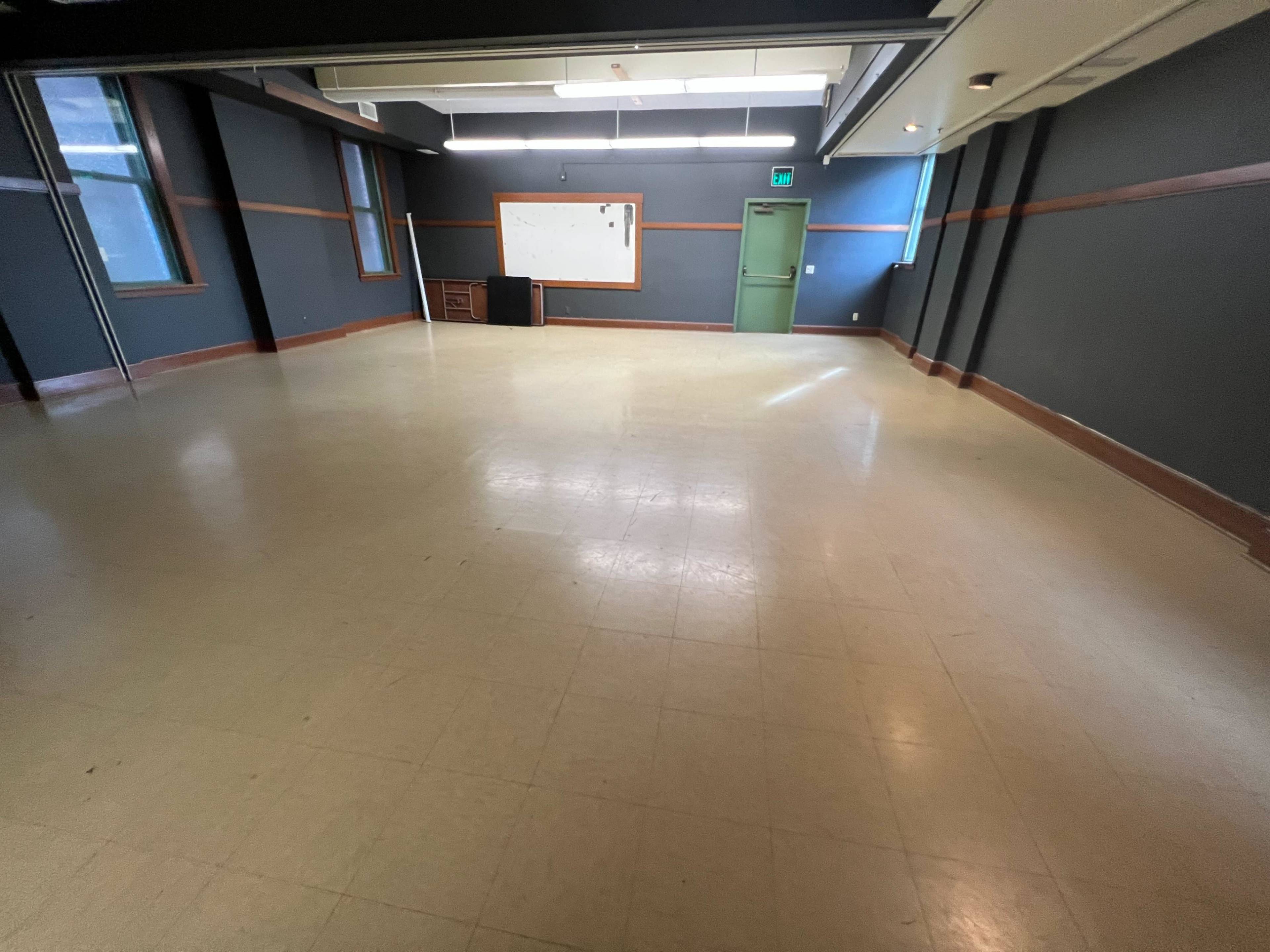 The image shows an empty classroom with a whiteboard, a desk in the corner, and green doors under overhead lights.