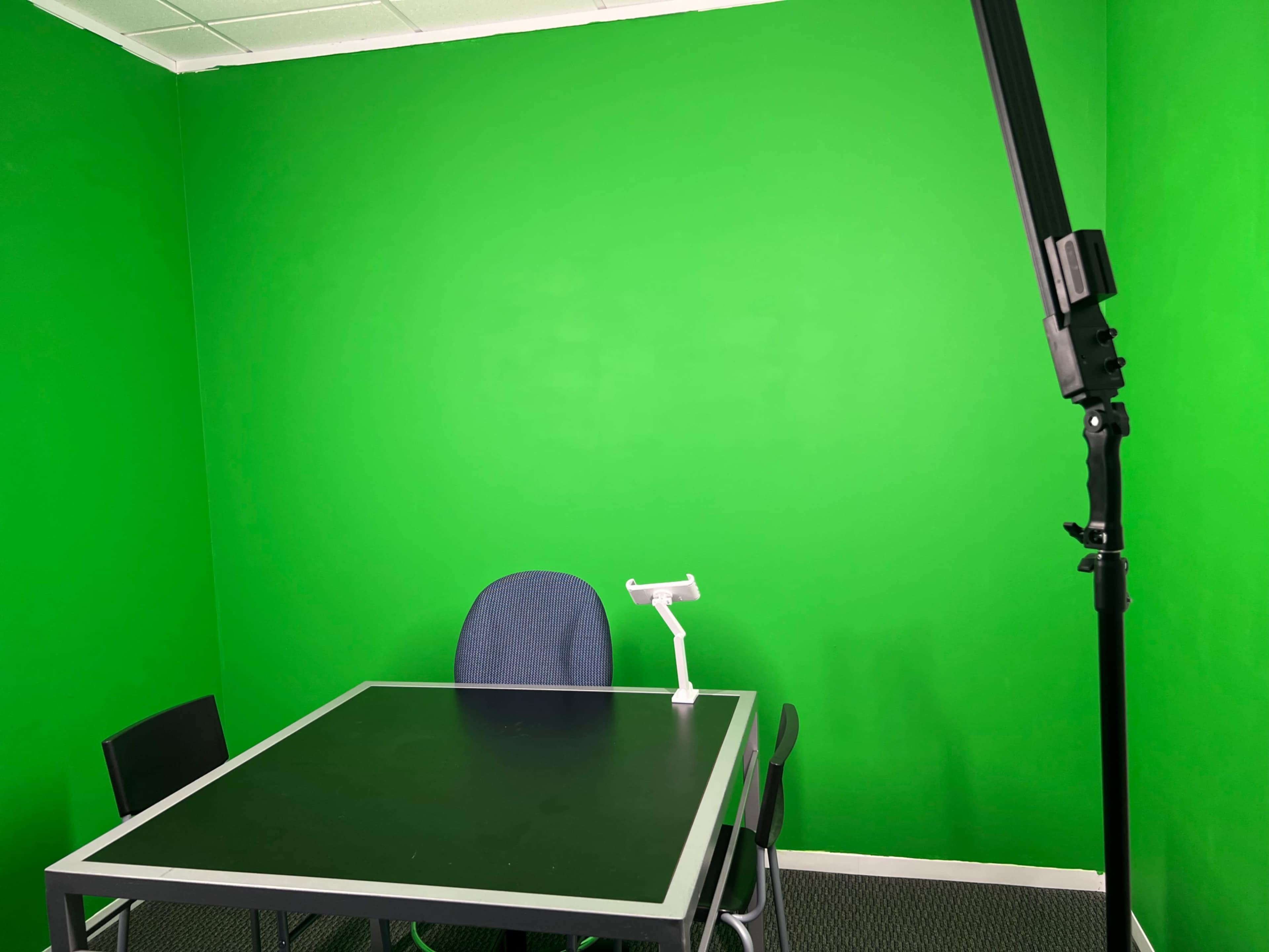 A green-walled room features a black table, a chair, and a lighting setup on a stand.