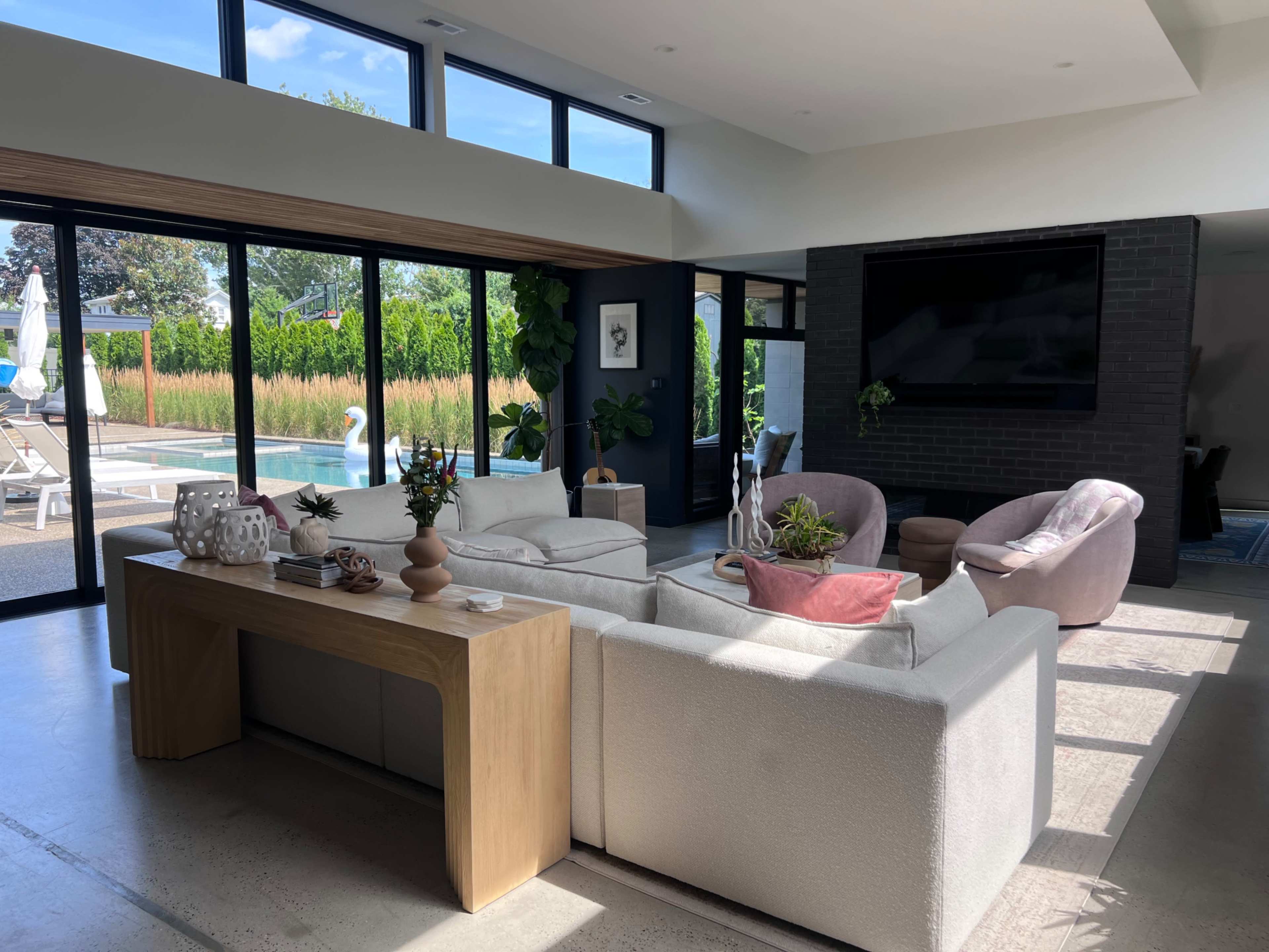 A modern living room features a light-colored sofa, a wooden coffee table, and large glass doors that open to a pool area surrounded by greenery.