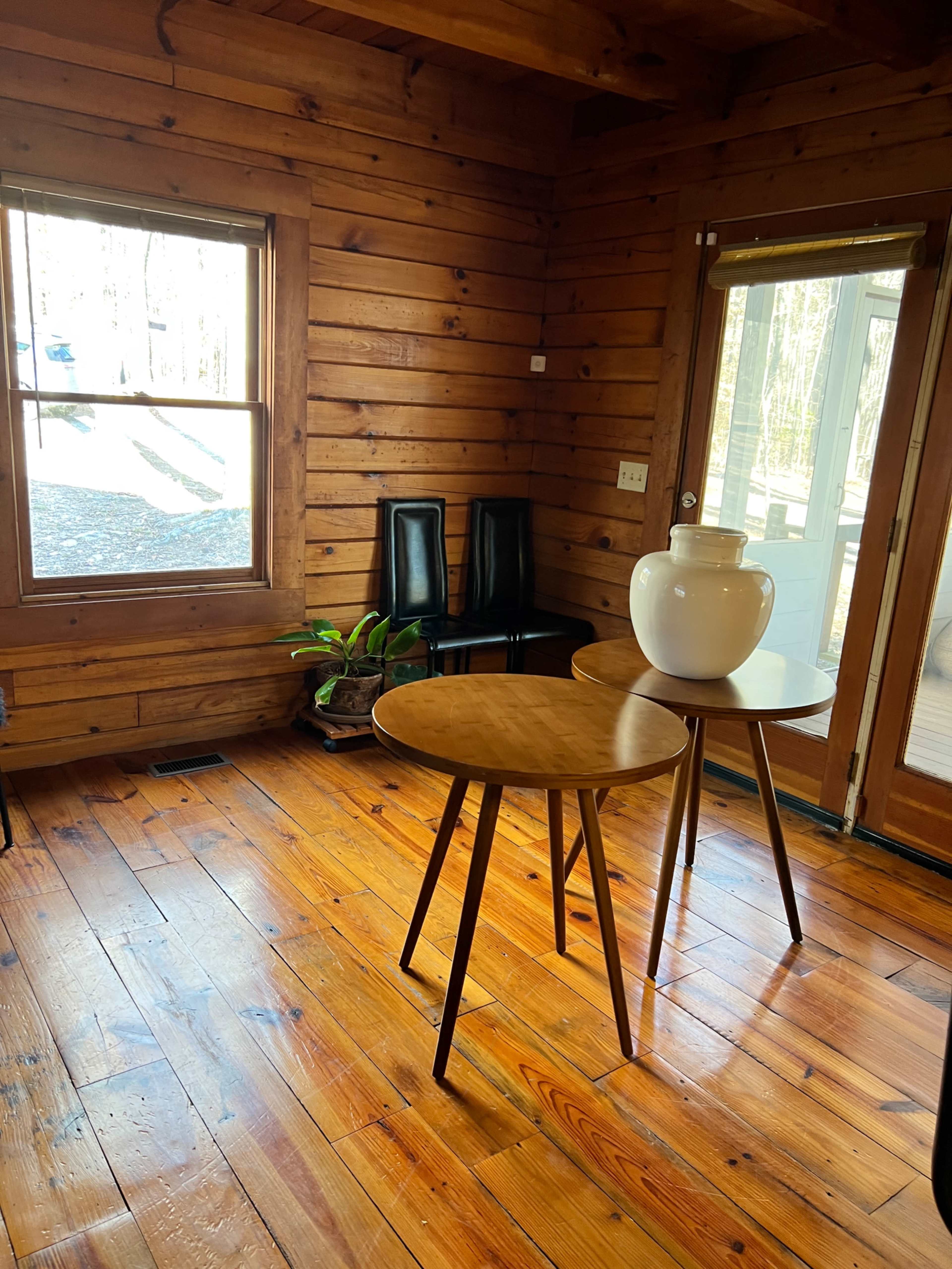 The interior of a wooden room features a round table with a large white vase on it, accompanied by two black chairs and a small plant near the window.