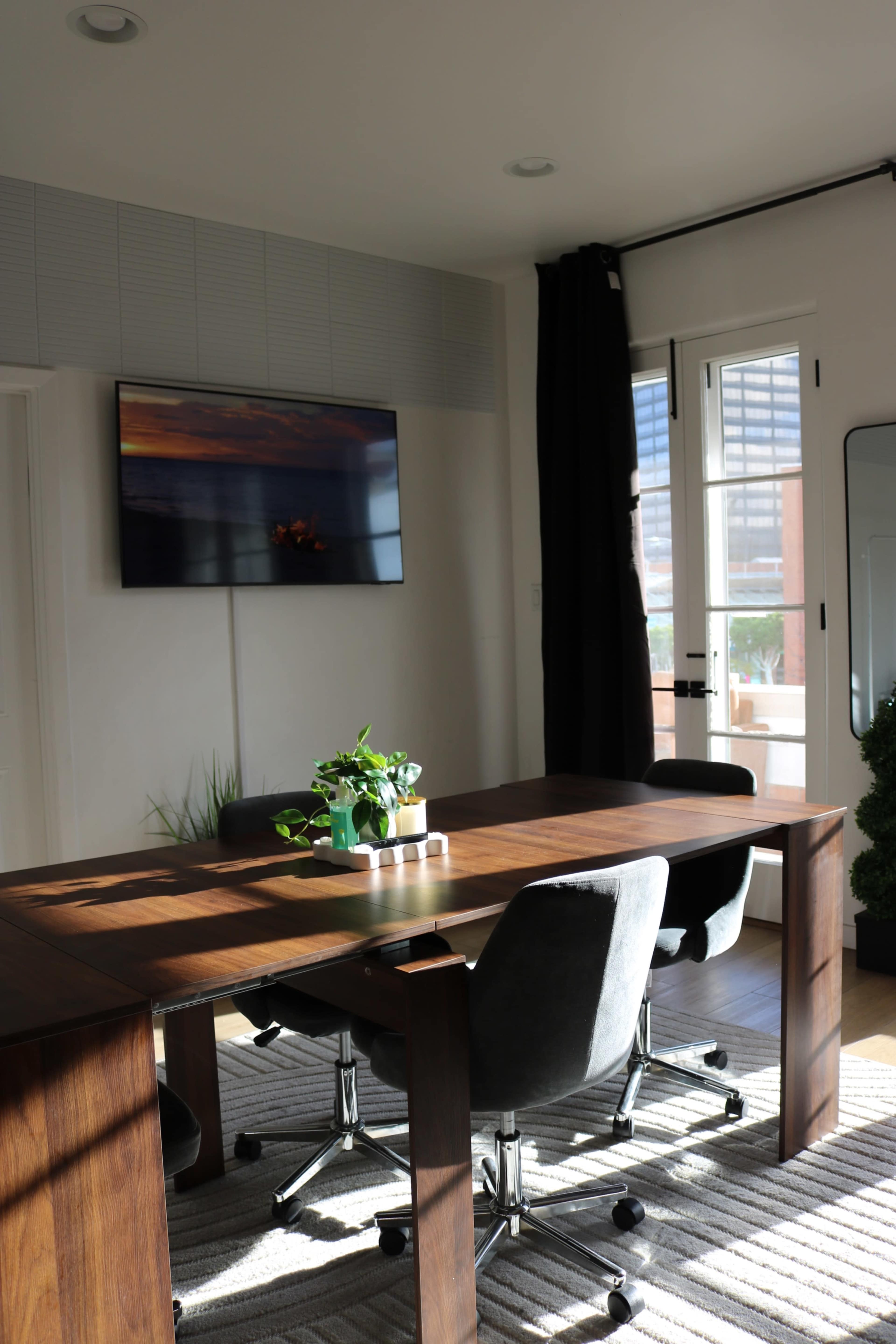 A modern conference room features a wooden table, black office chairs, a television mounted on the wall, and a large window with natural light.
