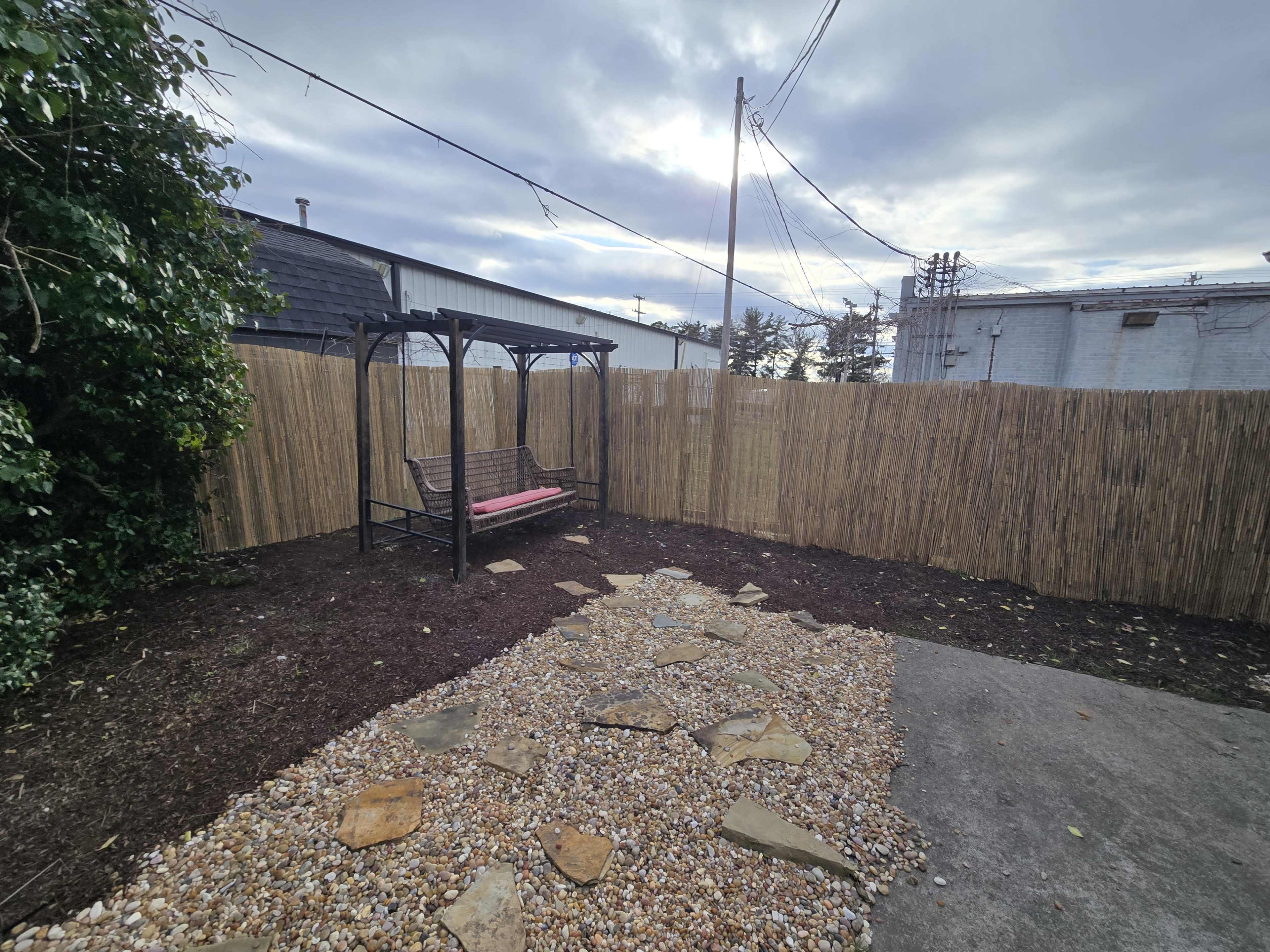 The image shows a fenced outdoor area with a gravel path leading to a swing bench under a pergola, surrounded by grass and wooden fencing.