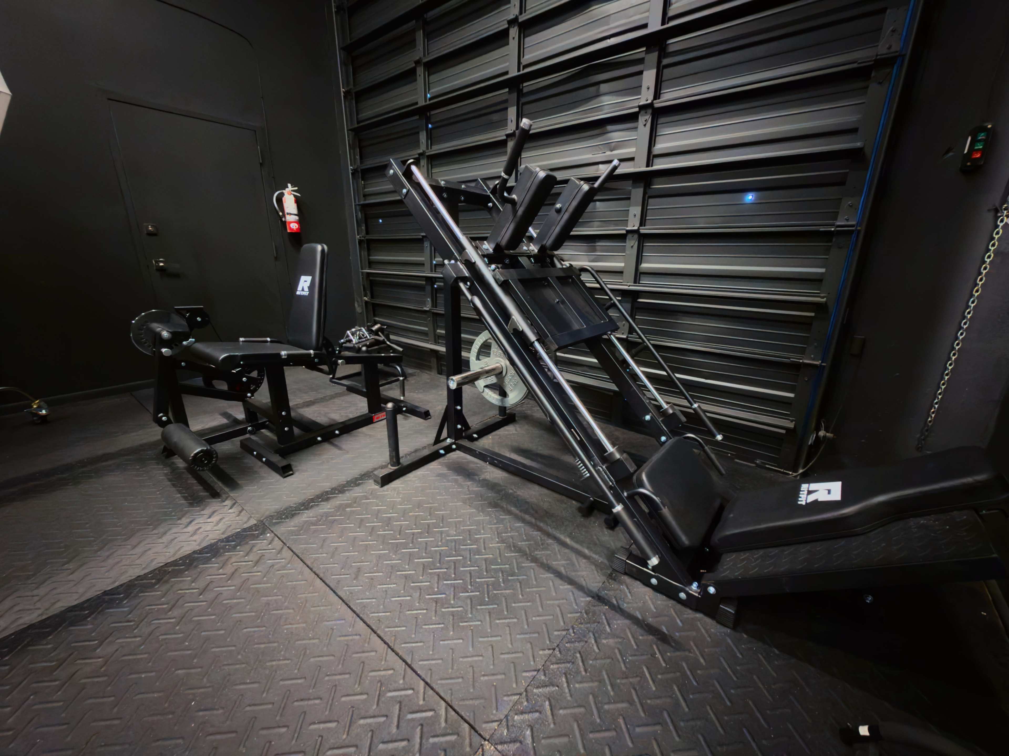 The image shows a gym facility with two pieces of weightlifting equipment: a leg press machine on the right and a seated leg curl machine on the left, set against a dark wall and floor.