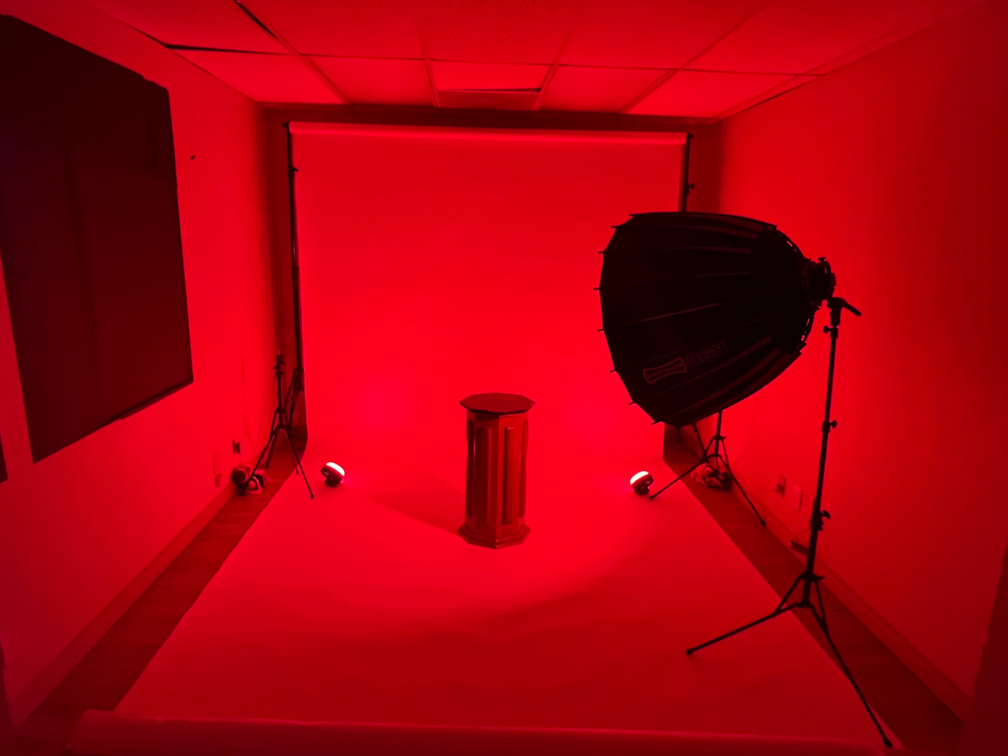 A photography studio with red lighting, featuring a pedestal in the center and a large softbox on the side.