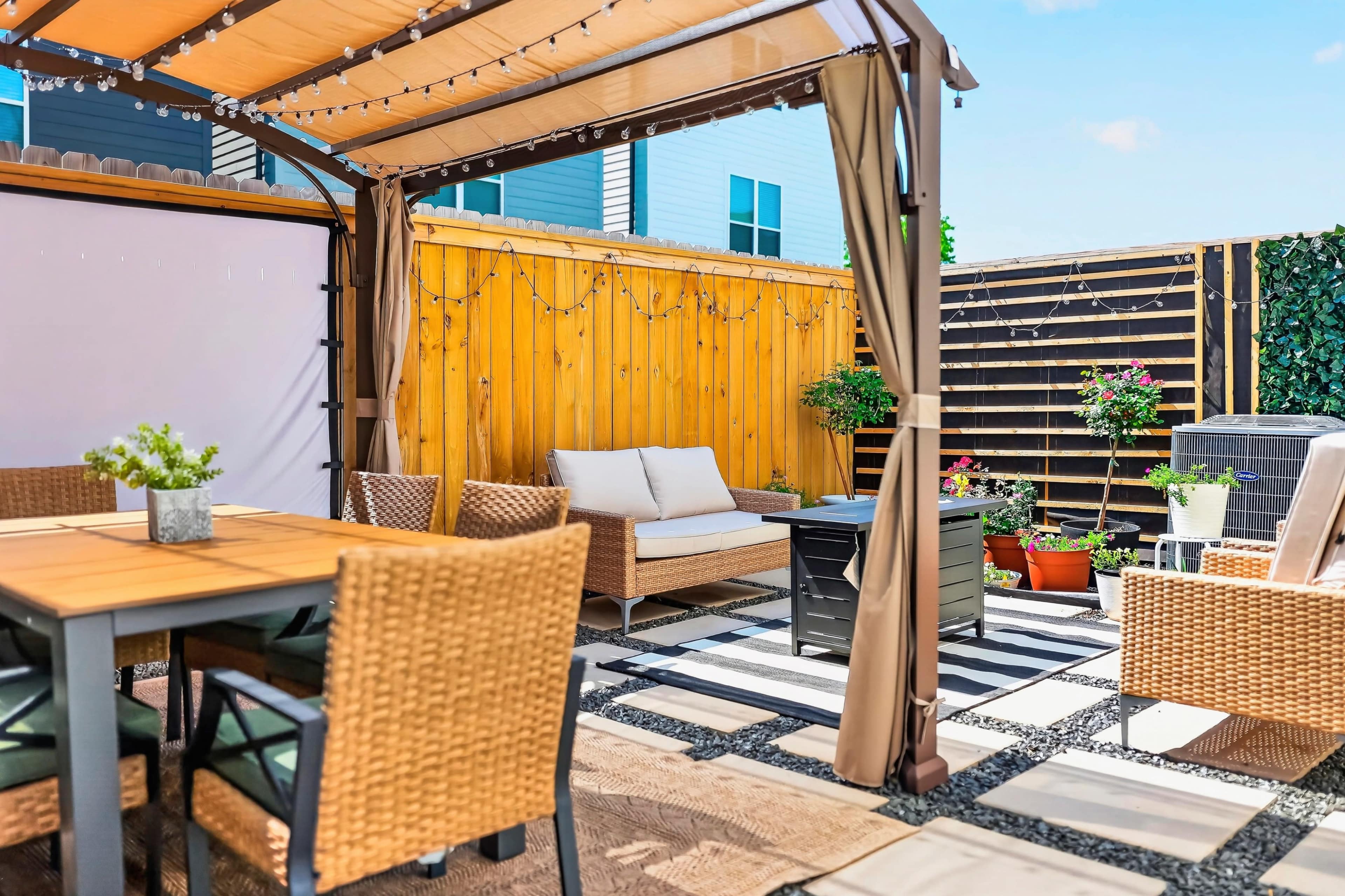 A patio area features a gazebo with seating, a table, and decorative plants against a backdrop of wooden fences and stone paving.
