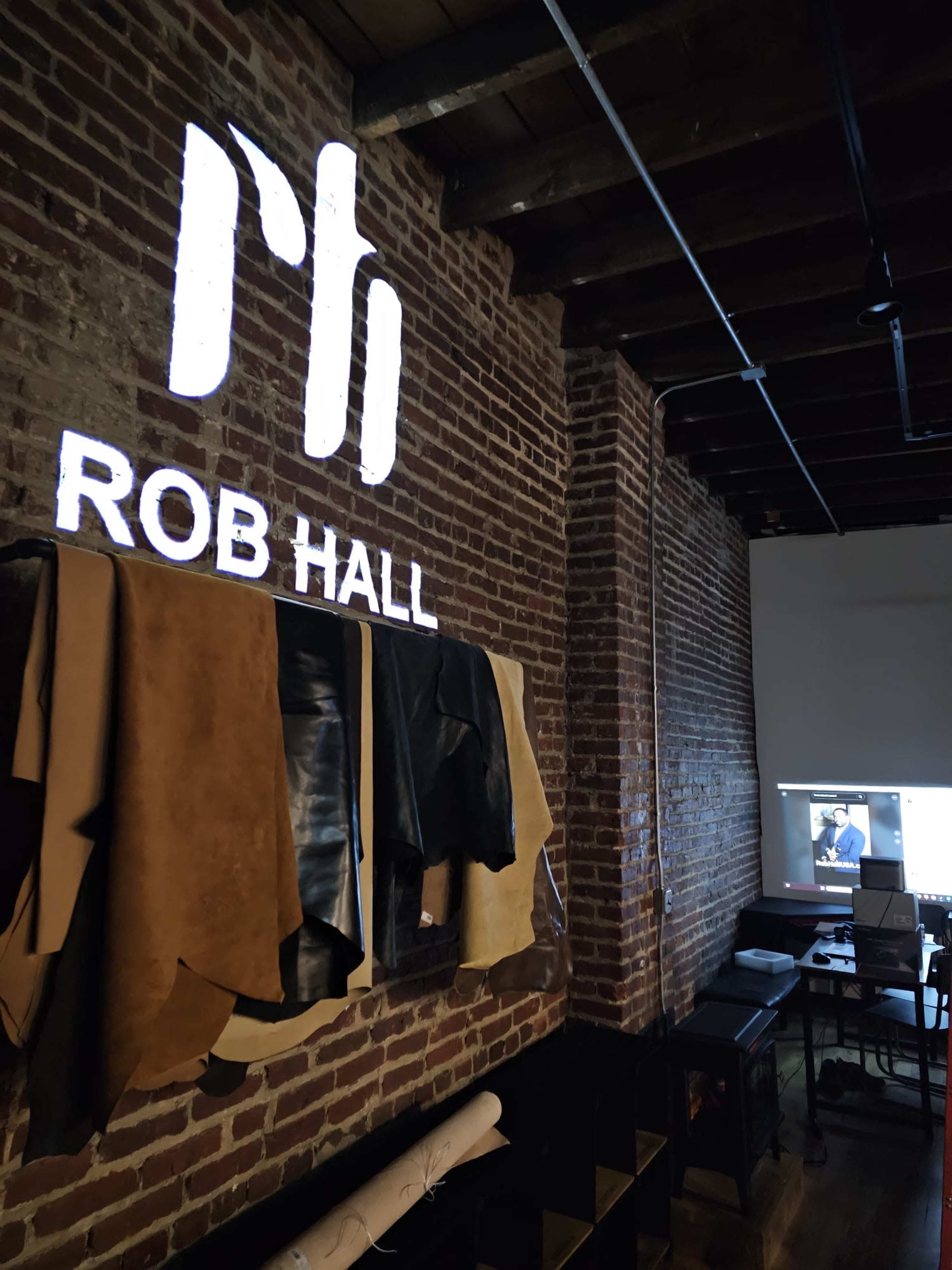 The image shows a brick wall featuring a logo and the name "ROB HALL" above hanging pieces of leather surrounded by a workspace with a projector showing an image.