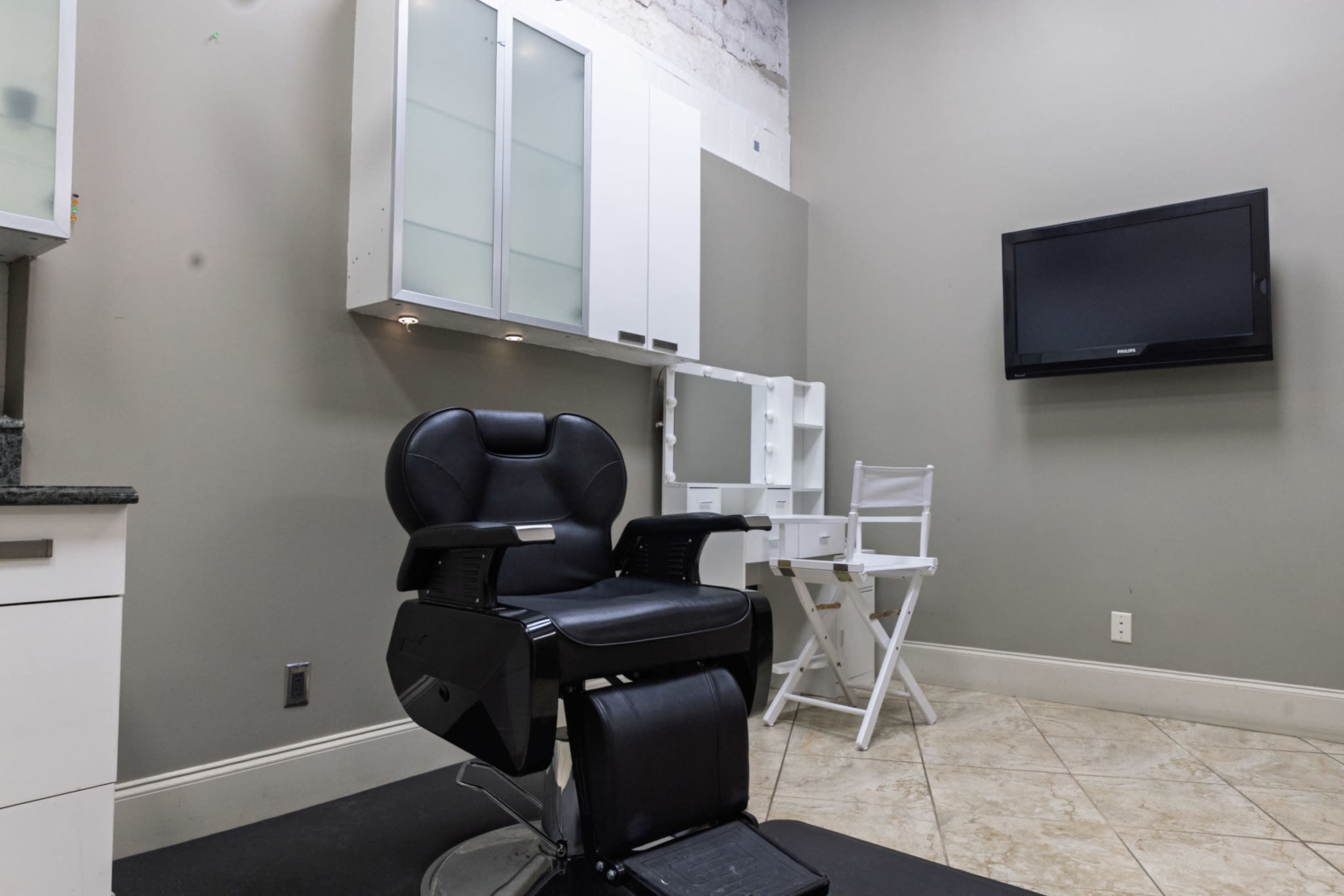 The image shows a minimalistic barber or salon setup with a black barber chair, a white vanity mirror with lights, and a television mounted on the wall.