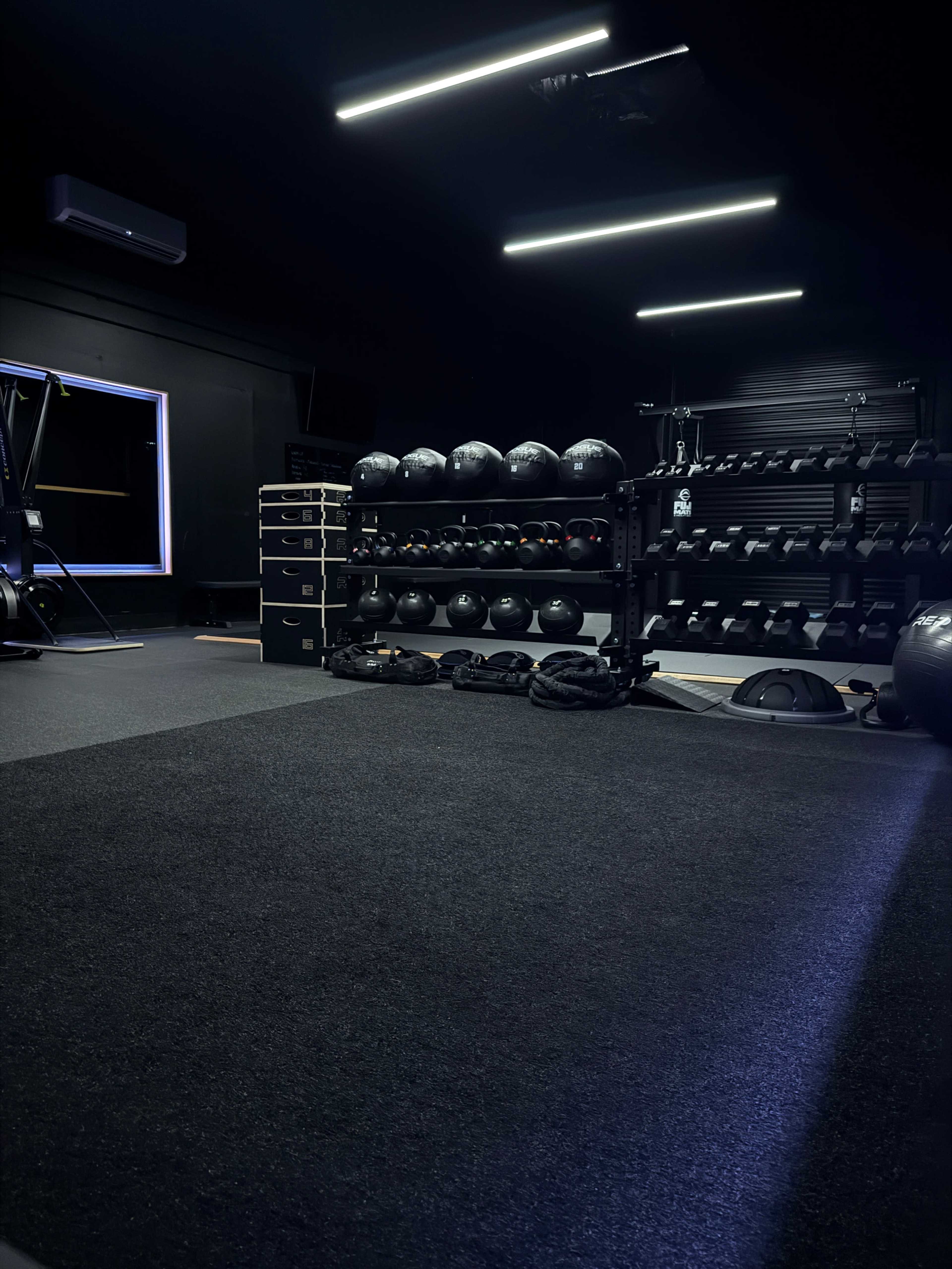 The image shows a dimly lit gym interior with a variety of weights and exercise equipment neatly organized on racks.