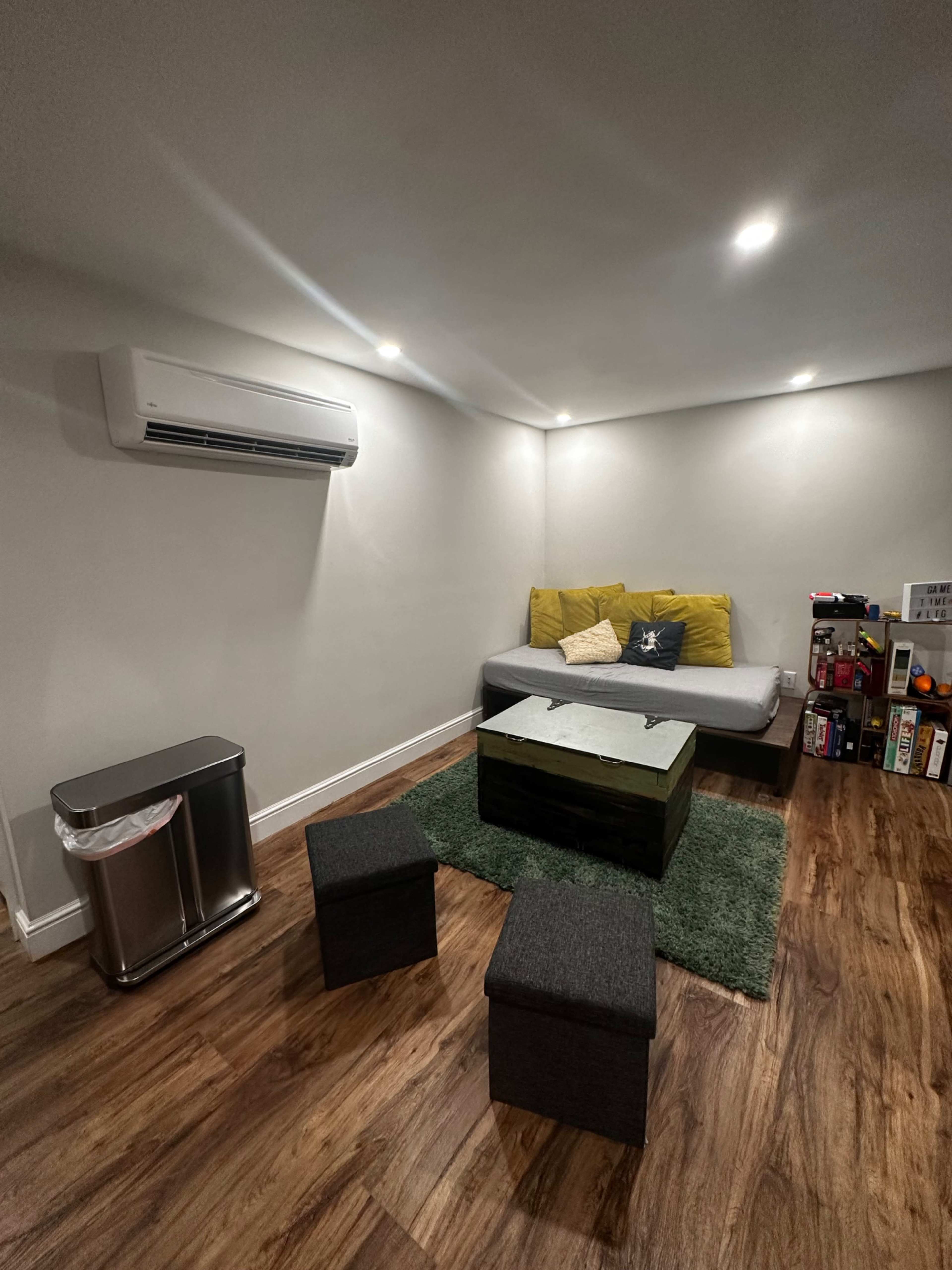 A cozy living space featuring a gray sofa with yellow throw pillows, a wooden coffee table, two black stools, a trash can, and a green rug on a hardwood floor, all under recessed lighting.