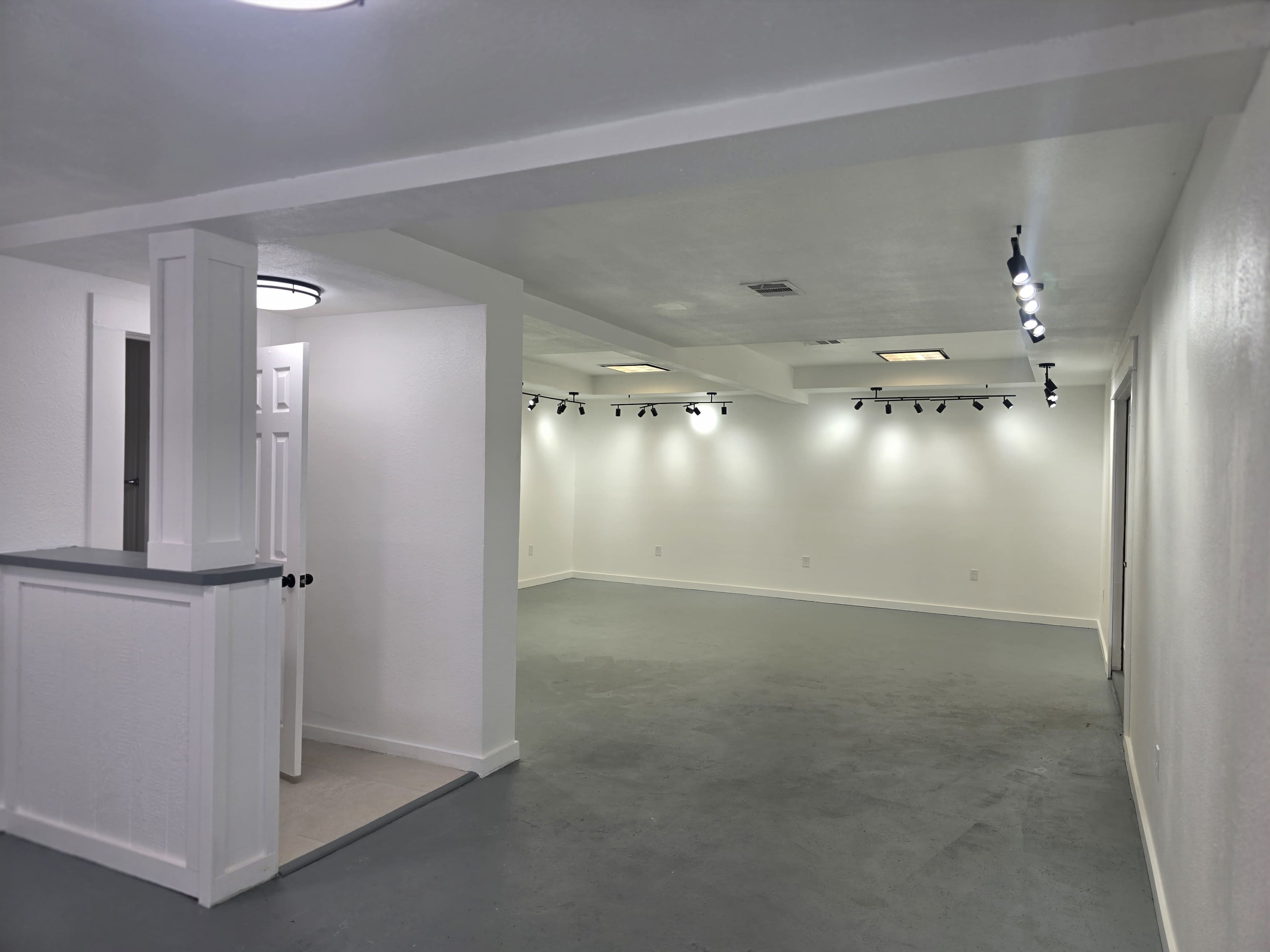 A spacious, empty room with concrete flooring and white walls, featuring track lighting along the ceiling.