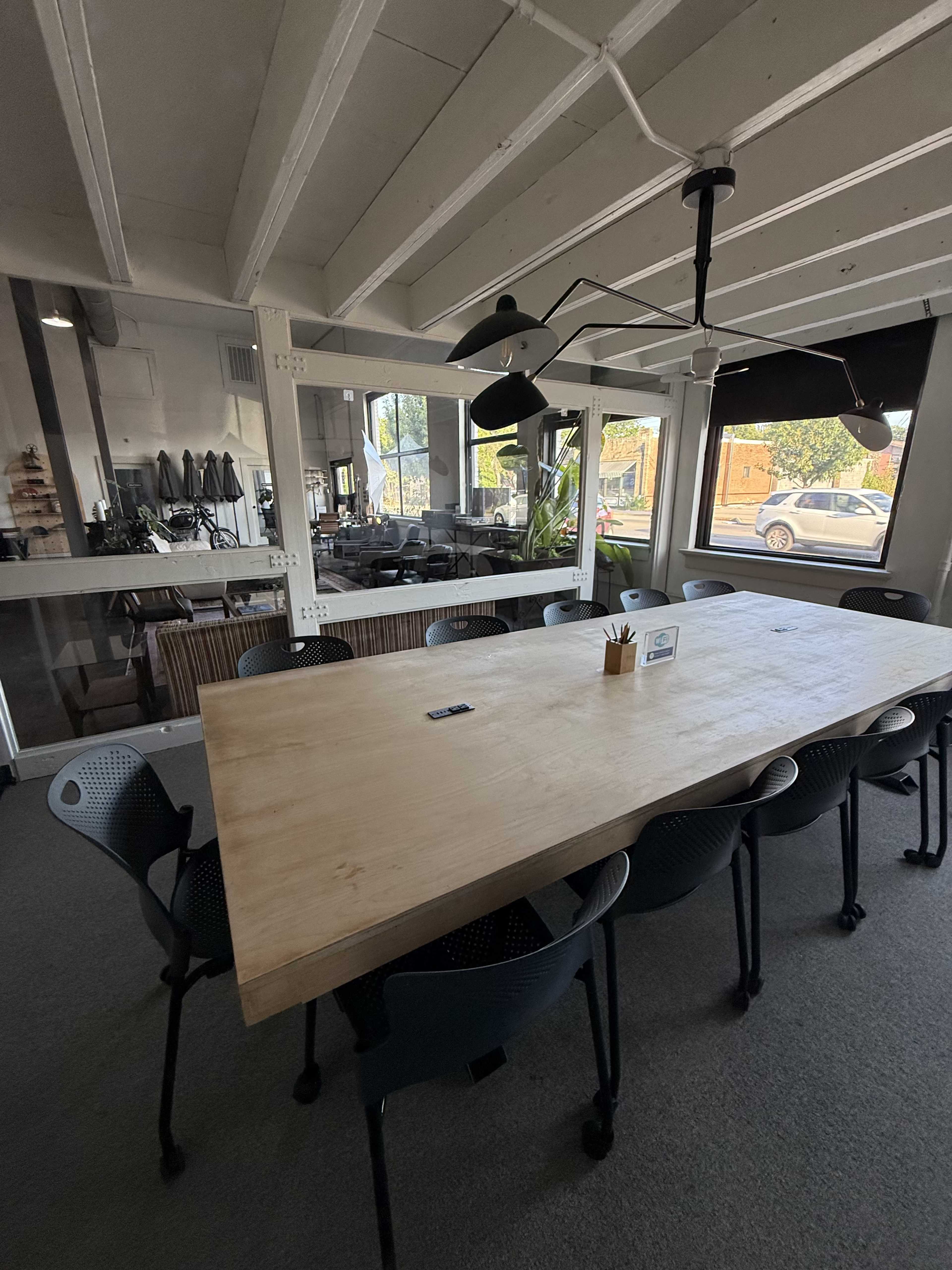 A large wooden table surrounded by black chairs sits in a well-lit modern meeting room with large windows.