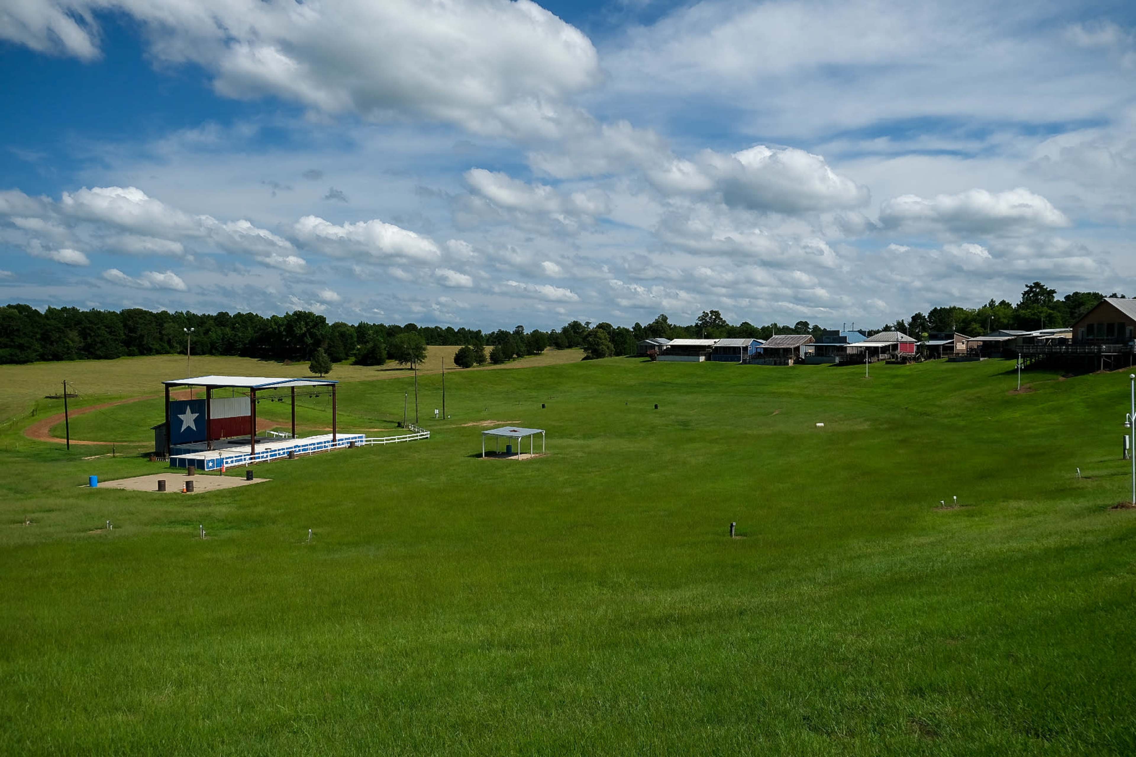 A grassy field stretches out with a stage featuring a Texas star on one side and a row of buildings on the other under a cloudy blue sky.