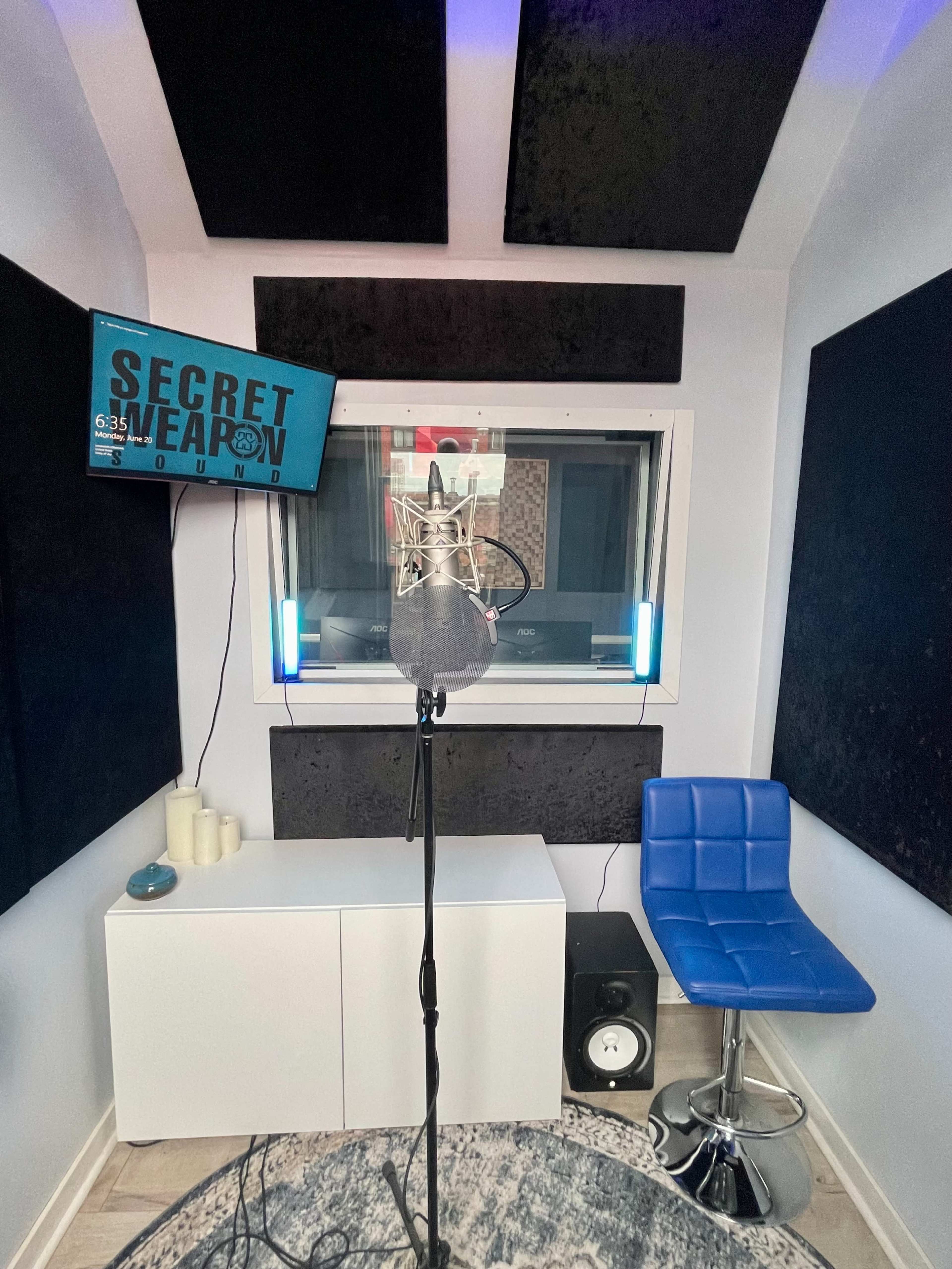 A recording studio booth with a microphone positioned in front of a soundproofed wall and a blue chair nearby.