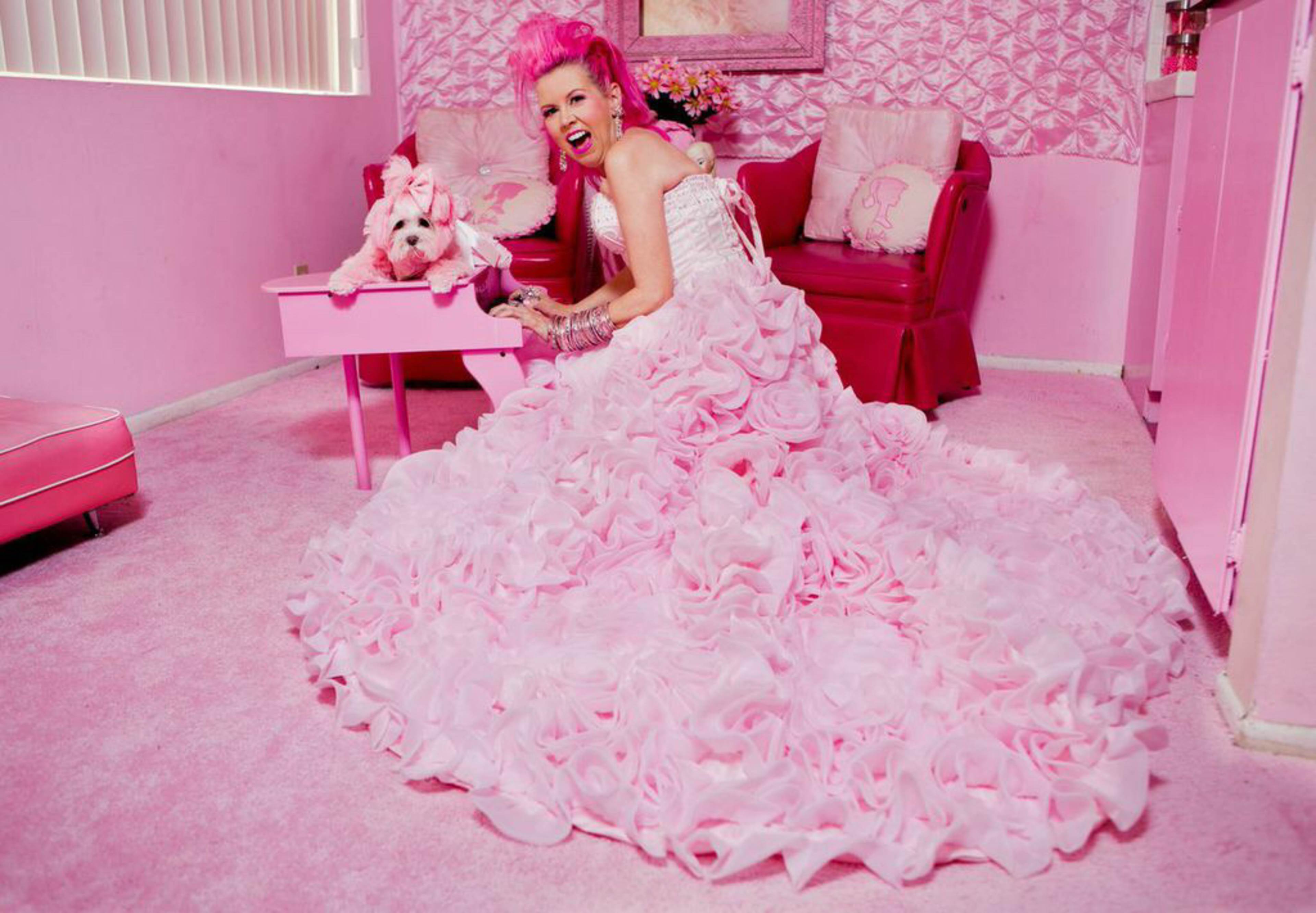 A woman in a voluminous pink gown poses in a pink room, holding a small dog beside a pink table.