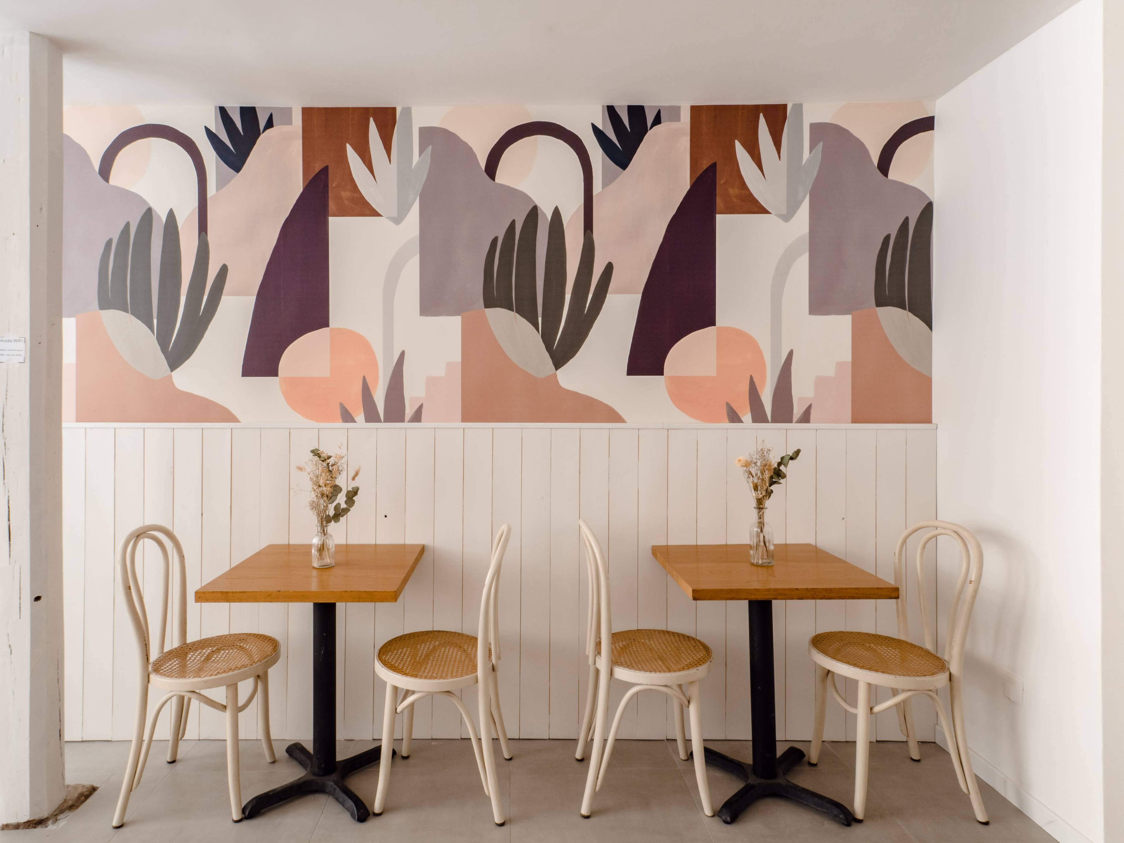The image shows a modern café interior with two wooden tables and four chairs, set against a patterned mural featuring abstract shapes and muted colors.