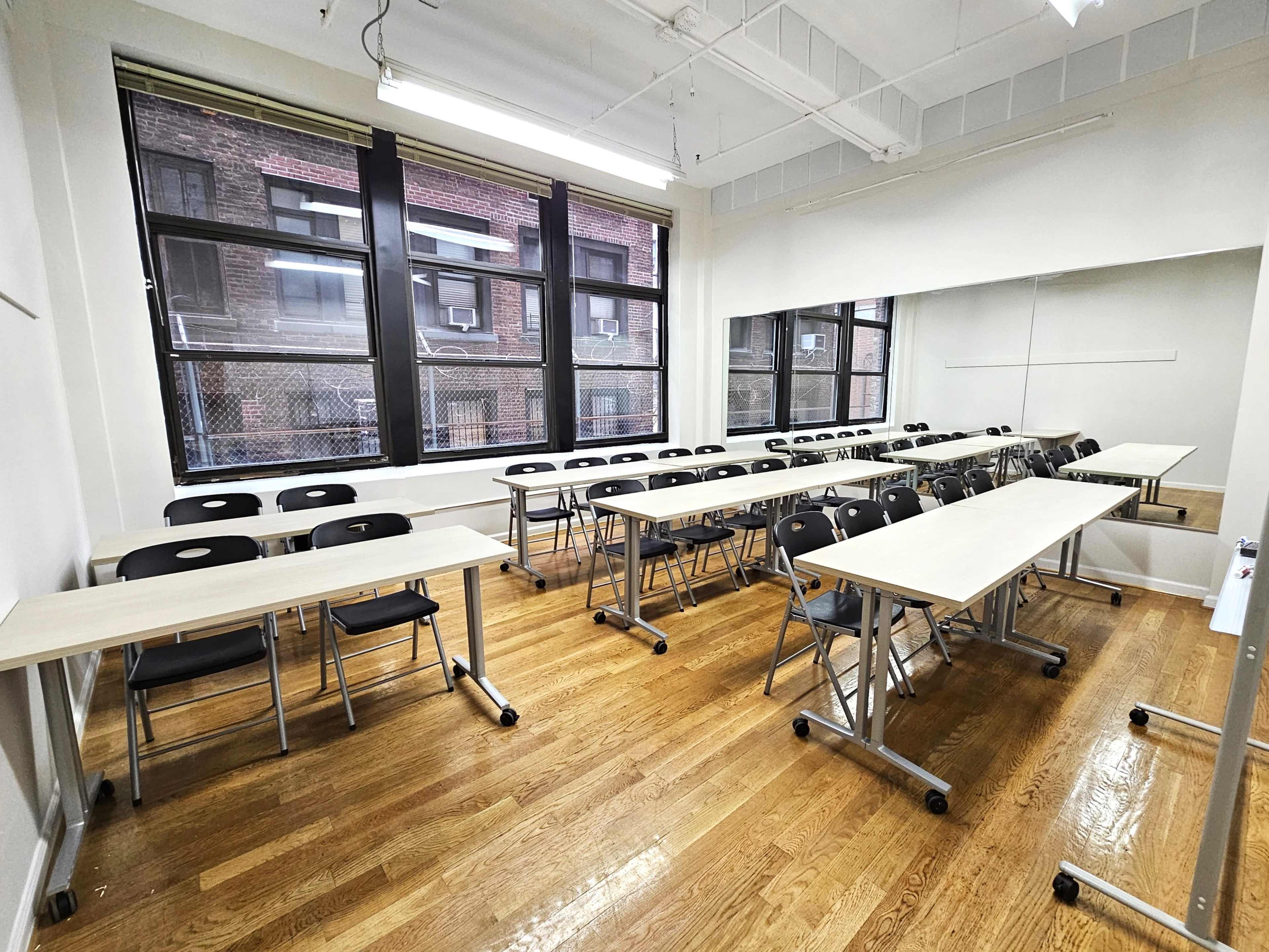 A classroom with several light-colored tables arranged in a U-shape and black chairs positioned around them, alongside large windows that overlook a brick building.