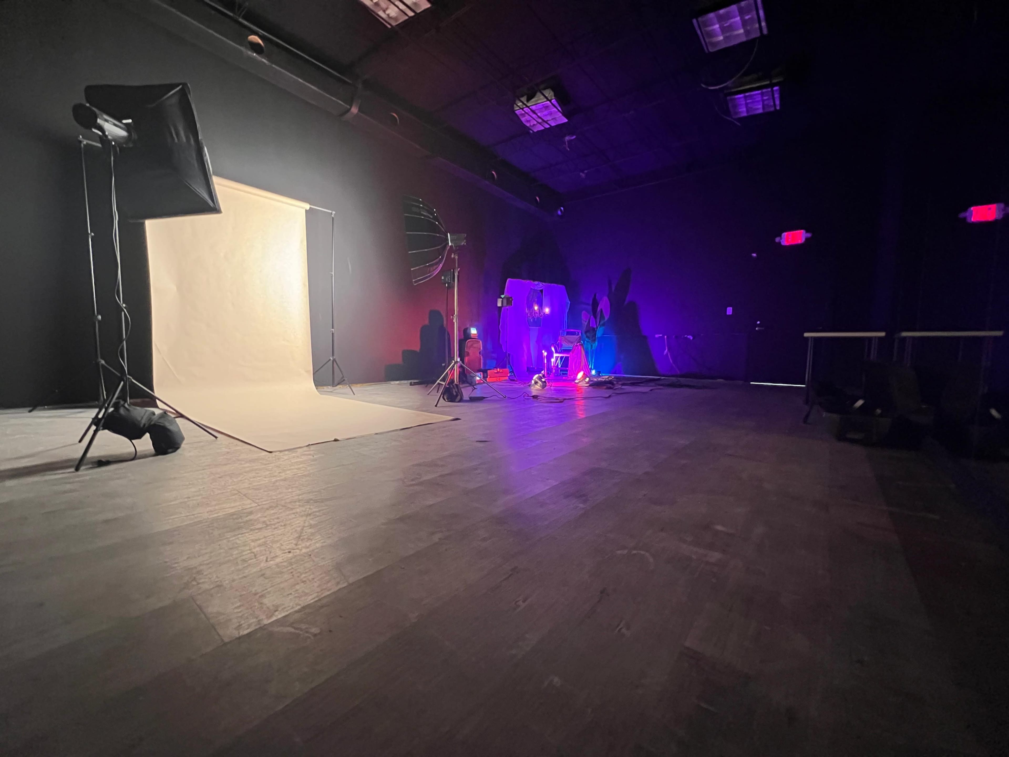 A photography studio features a light backdrop illuminated by various lighting equipment and props set against dark walls and a wooden floor.