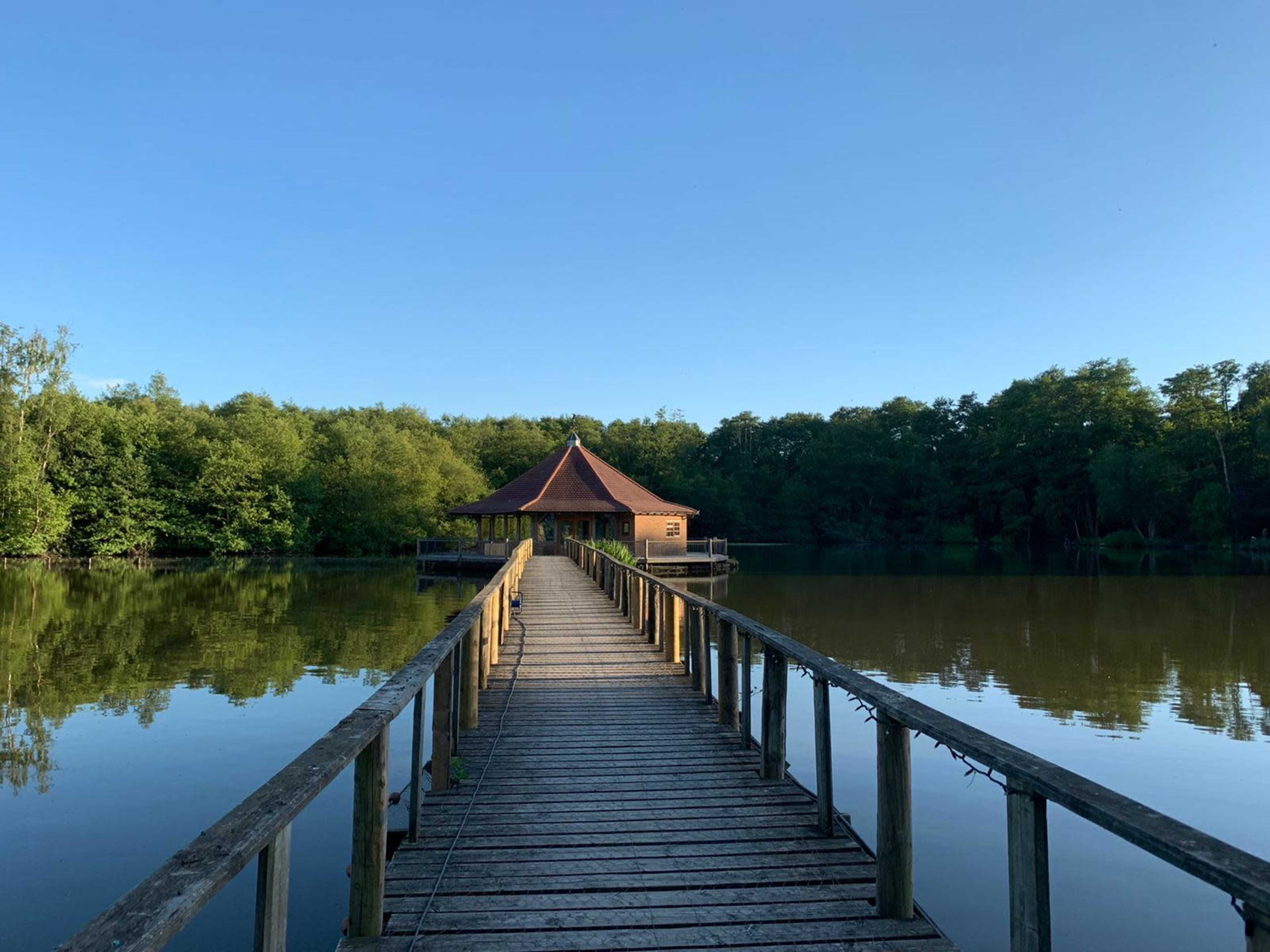 A wooden pier leads to a gazebo-style structure on the edge of a calm lake, surrounded by trees.