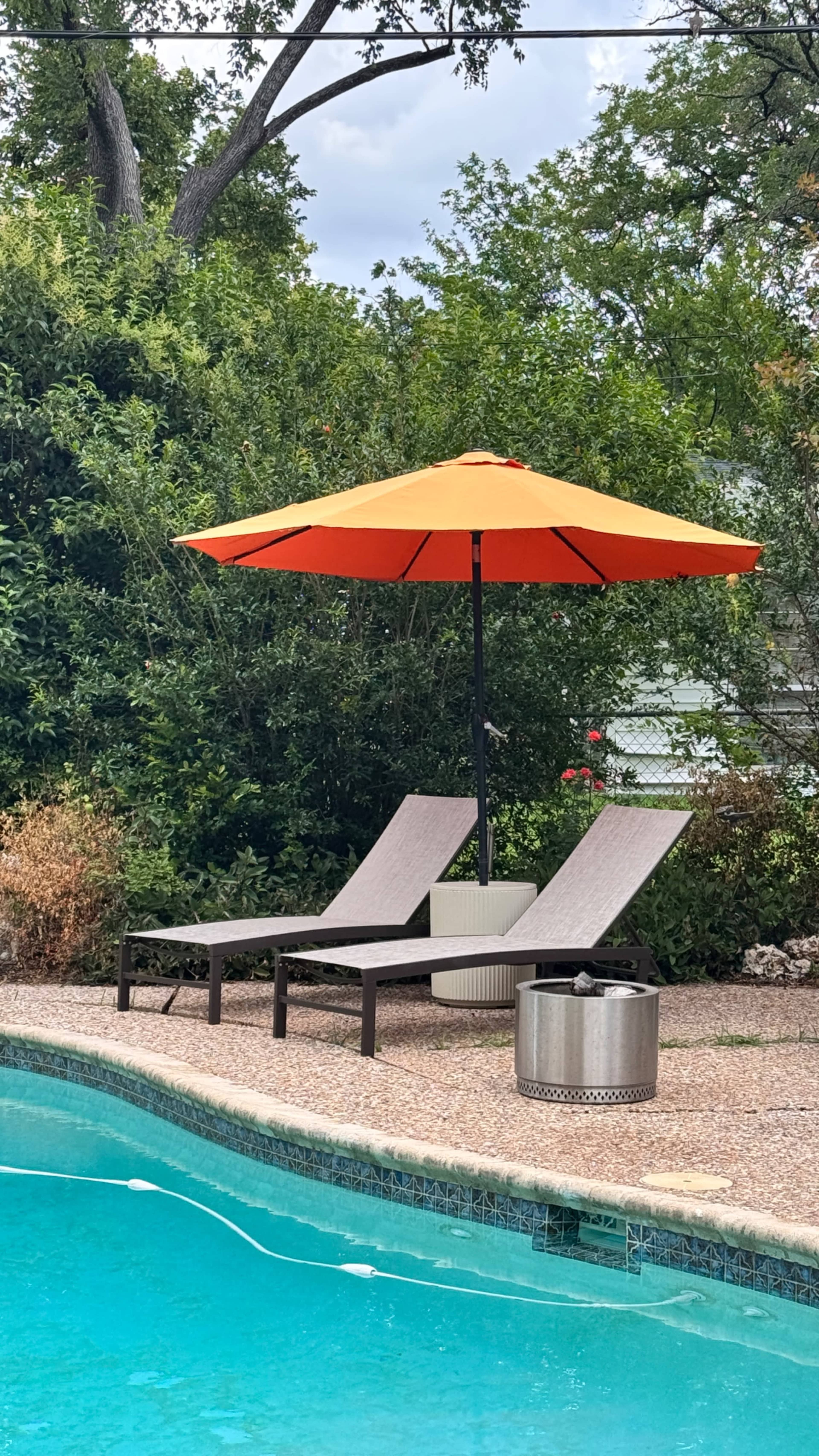The image shows two lounge chairs beneath an orange umbrella next to a swimming pool, with a metal fire pit nearby and greenery in the background.