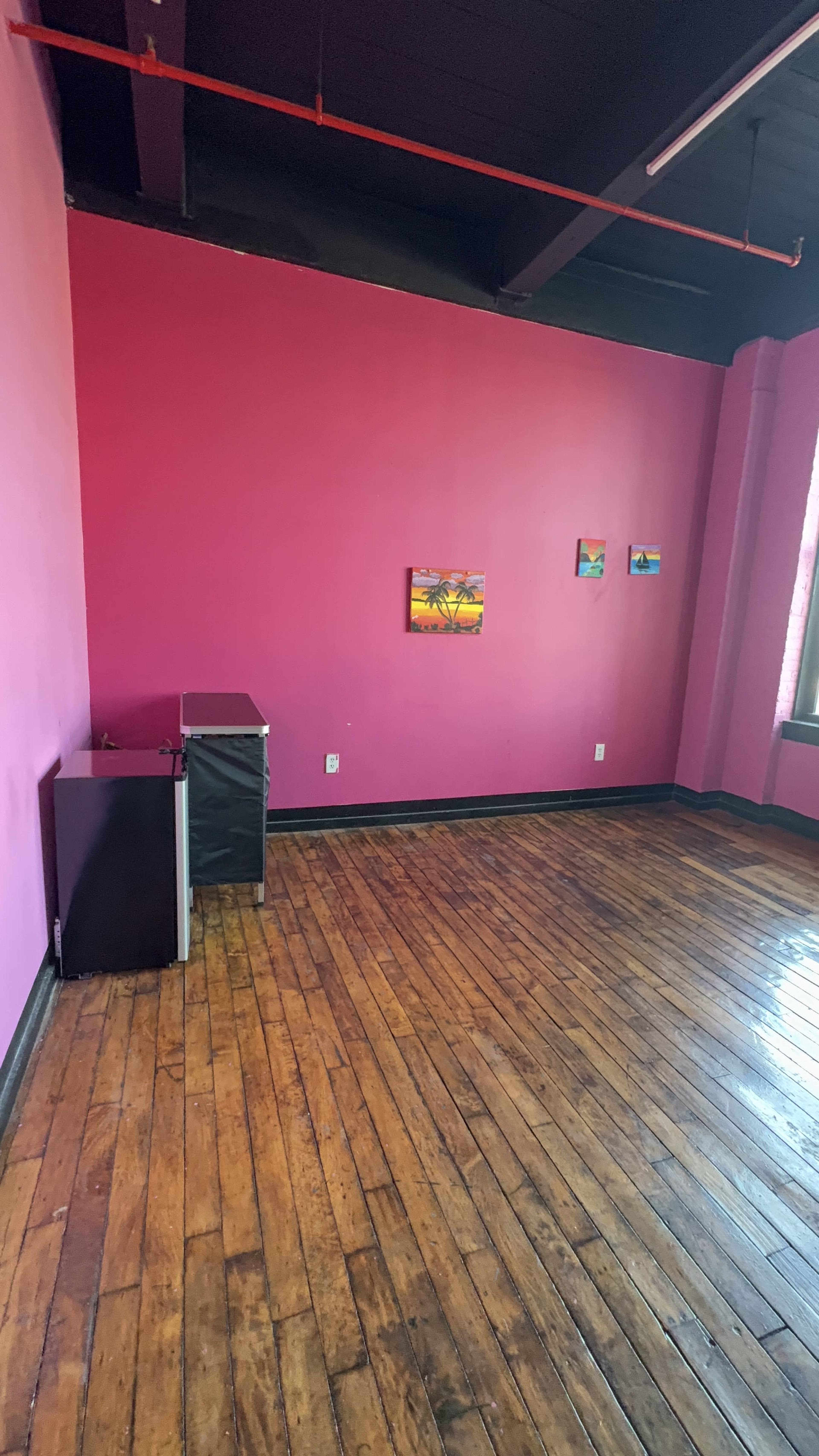 The image shows a brightly colored pink room with wooden flooring, featuring three small pieces of artwork on the wall and a small desk or storage unit in one corner.