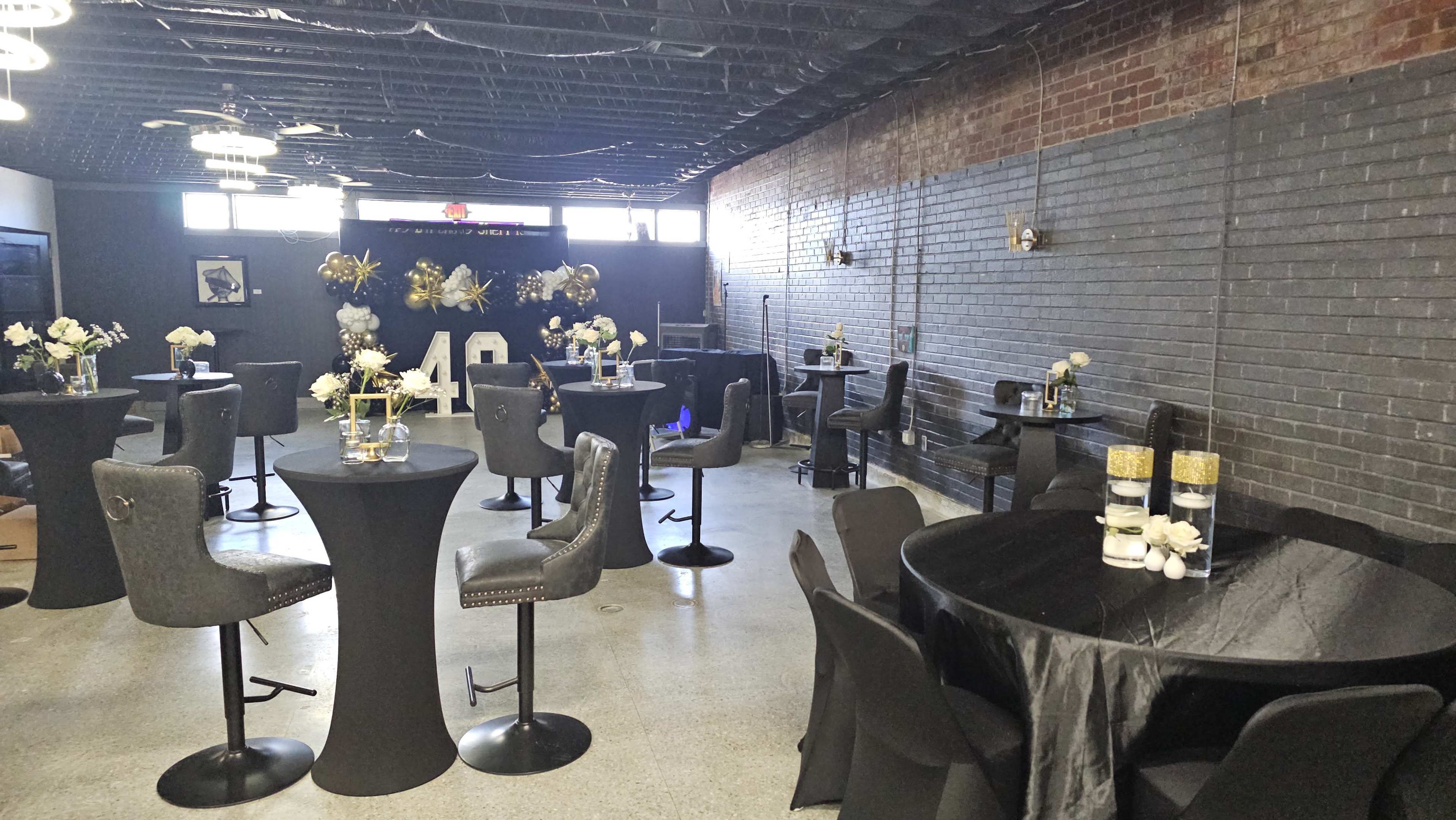 The image shows a decorated event space with black and gold tables, chairs, and floral arrangements.