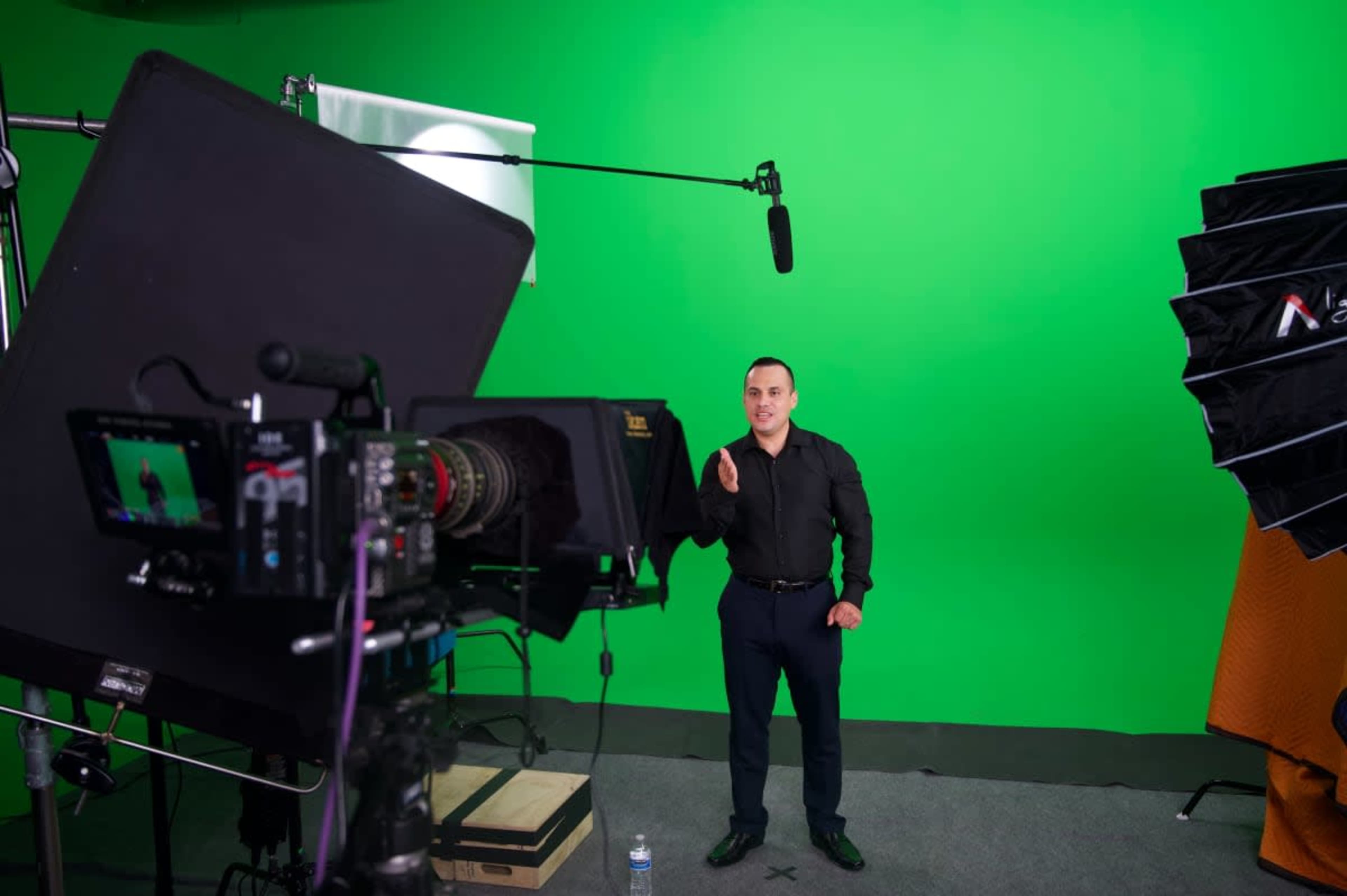 A man stands in front of a green screen, gesturing while multiple cameras and lighting equipment surround him.