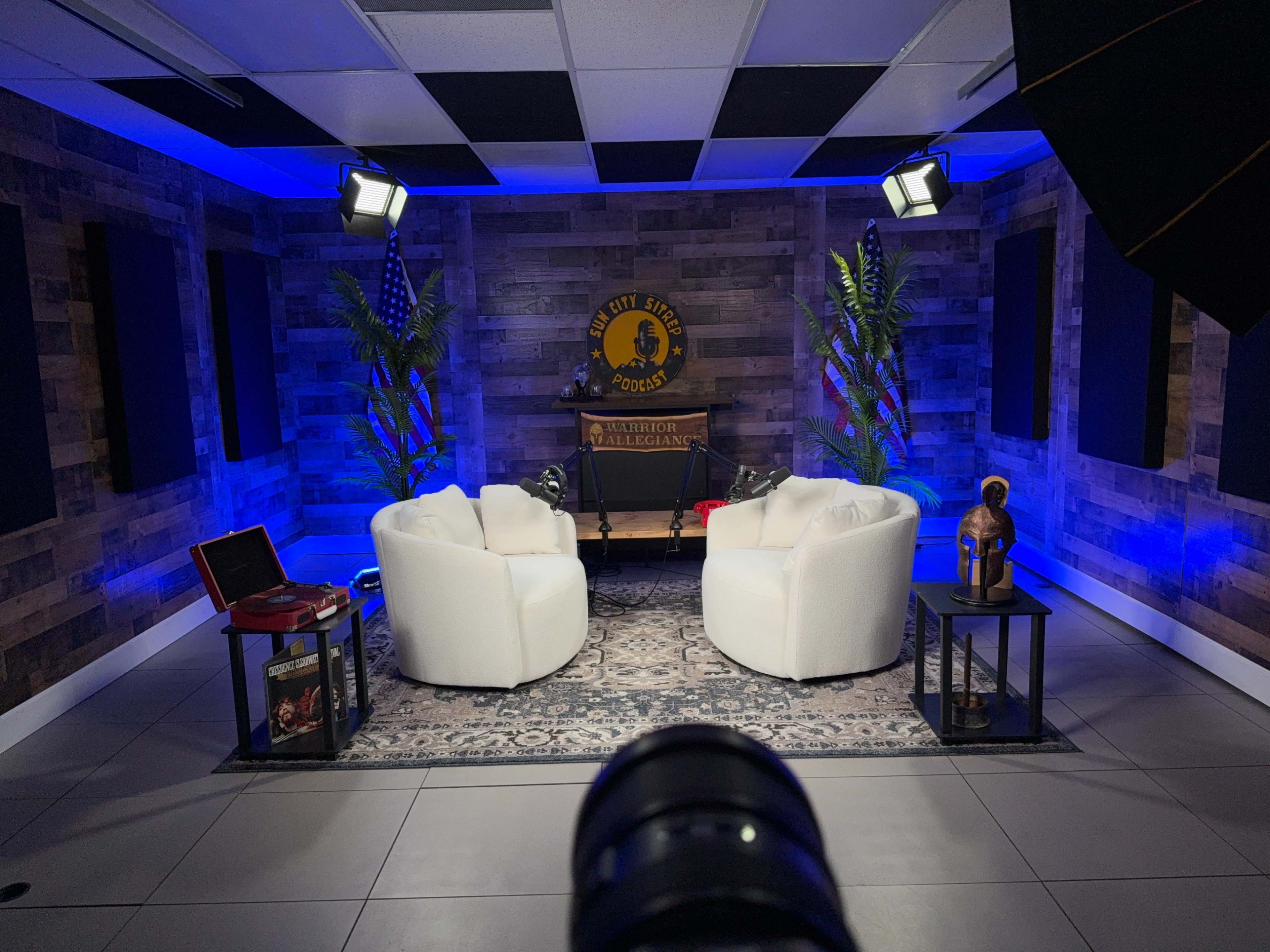 The image shows a podcast studio featuring two white chairs, a table in between, and decorative plants, all set against a wooden wall and illuminated by blue lighting.
