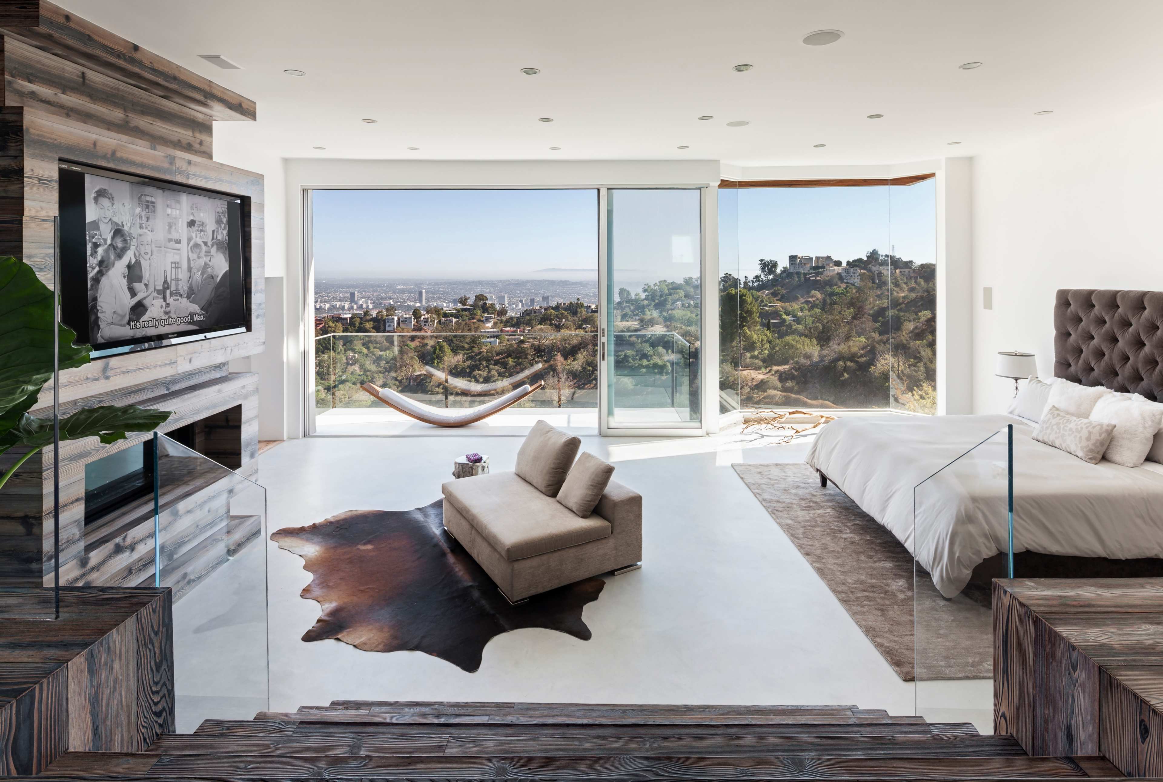 A modern bedroom with a large window overlooking a cityscape, featuring a television mounted on a wall, a bed, a lounge chair, and a hammock visible outside.