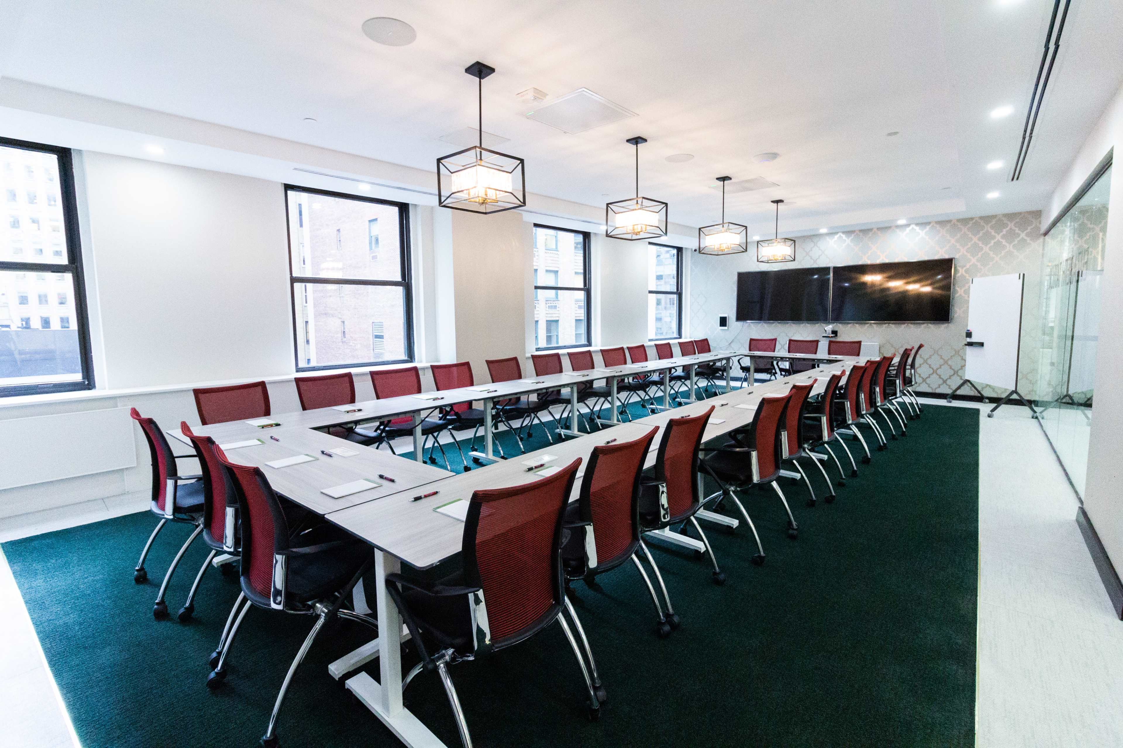A modern conference room features a long rectangular table surrounded by red chairs, with large windows and overhead lighting.