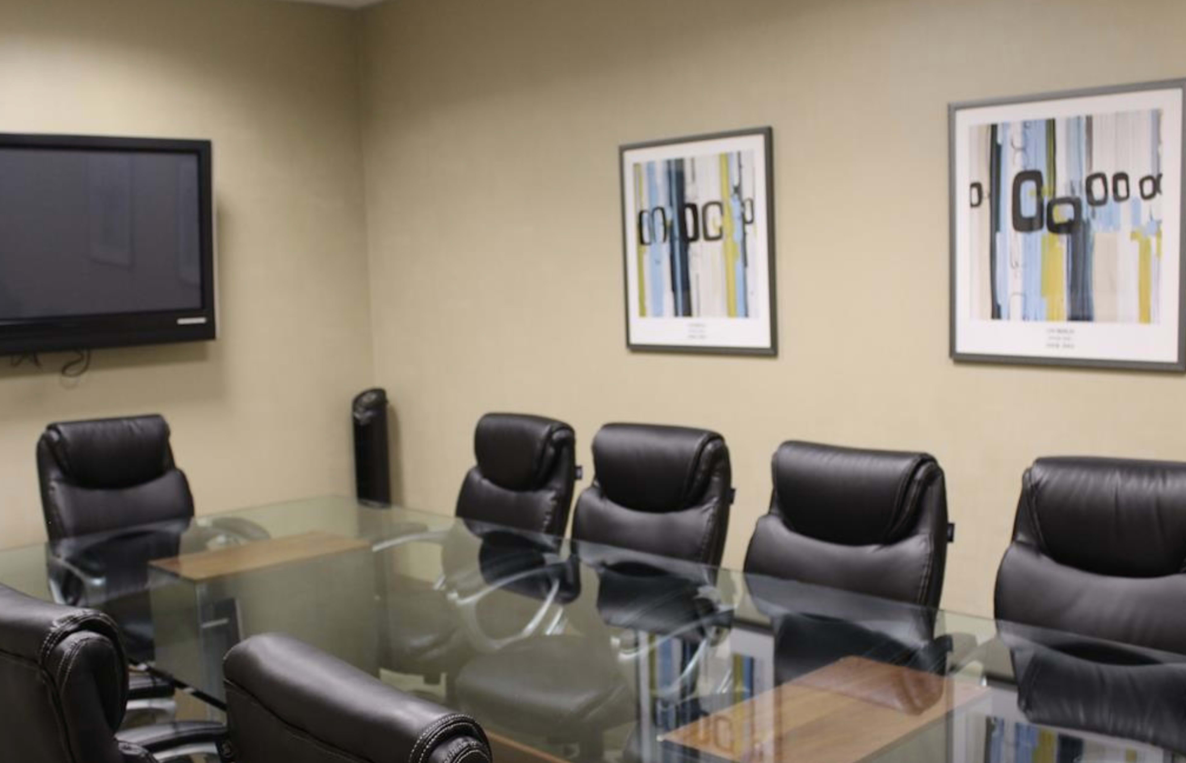 A conference room features a long glass table surrounded by black leather chairs, with two framed artworks on the wall and a TV mounted in the corner.