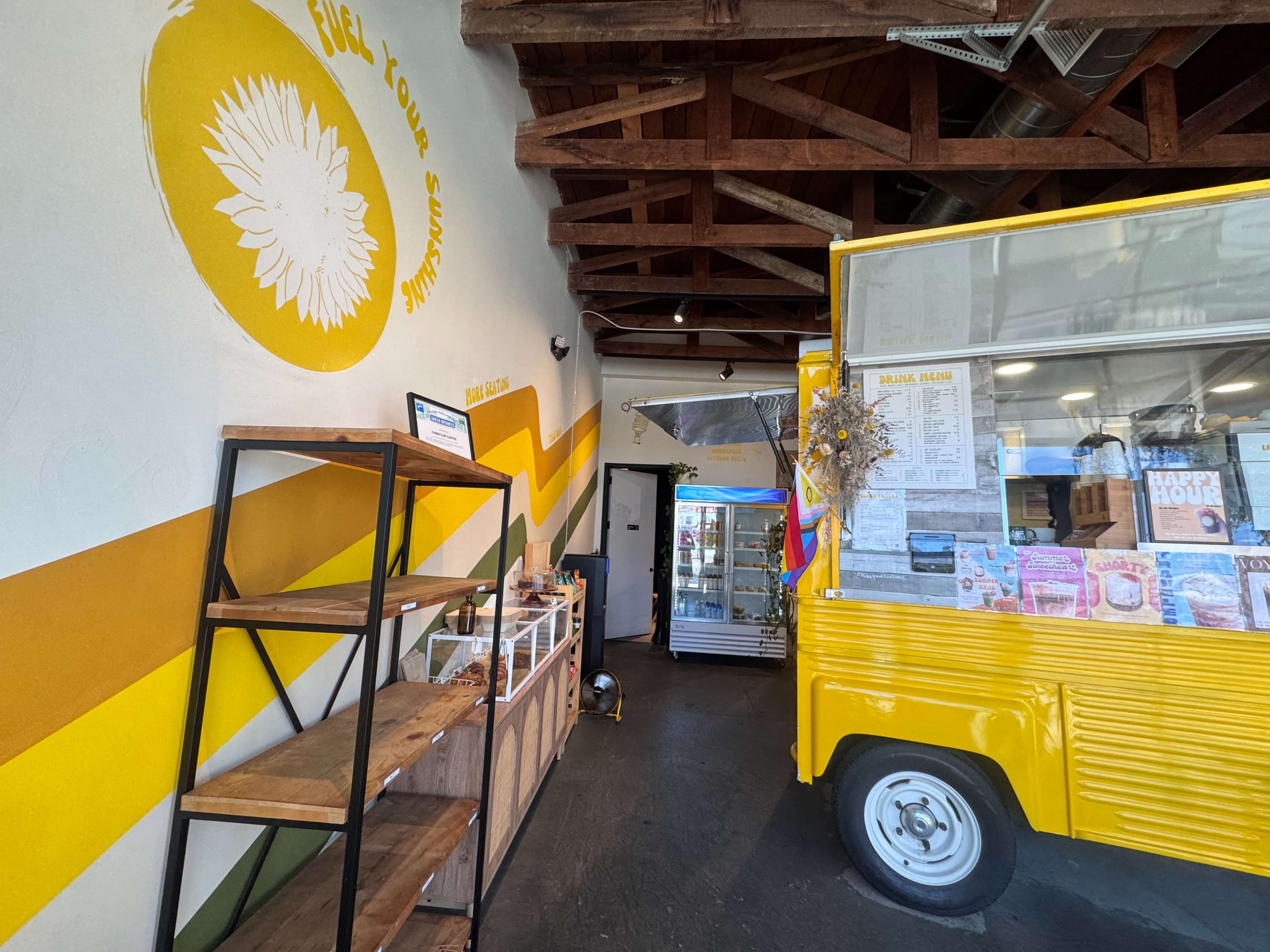 The image shows the interior of a small, brightly colored café with a wooden shelf displaying items and a food truck-style counter at one end.