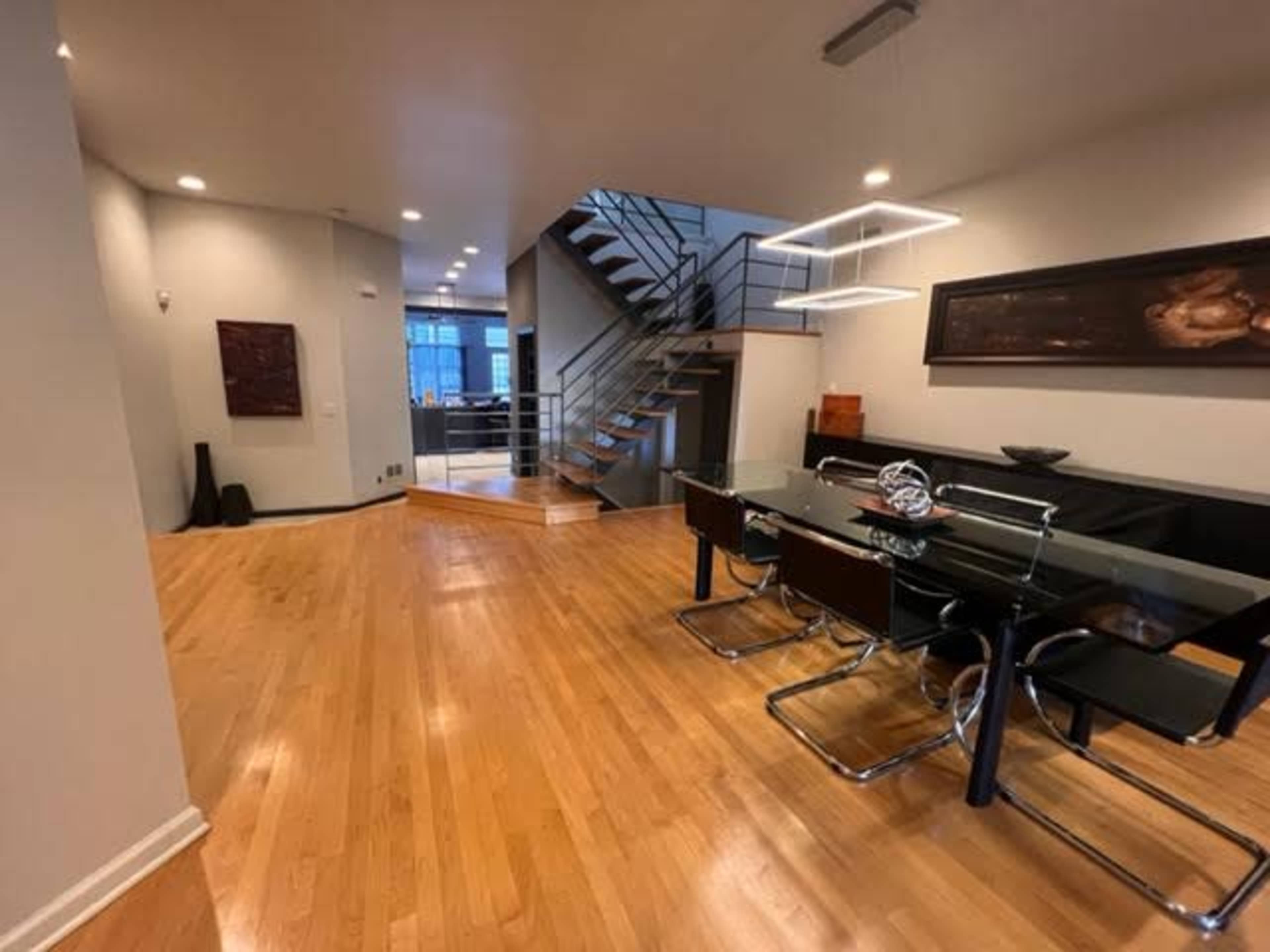 A modern dining area with a glass table and black chairs, positioned next to a staircase leading to an upper level in a well-lit, spacious room with wooden floors.