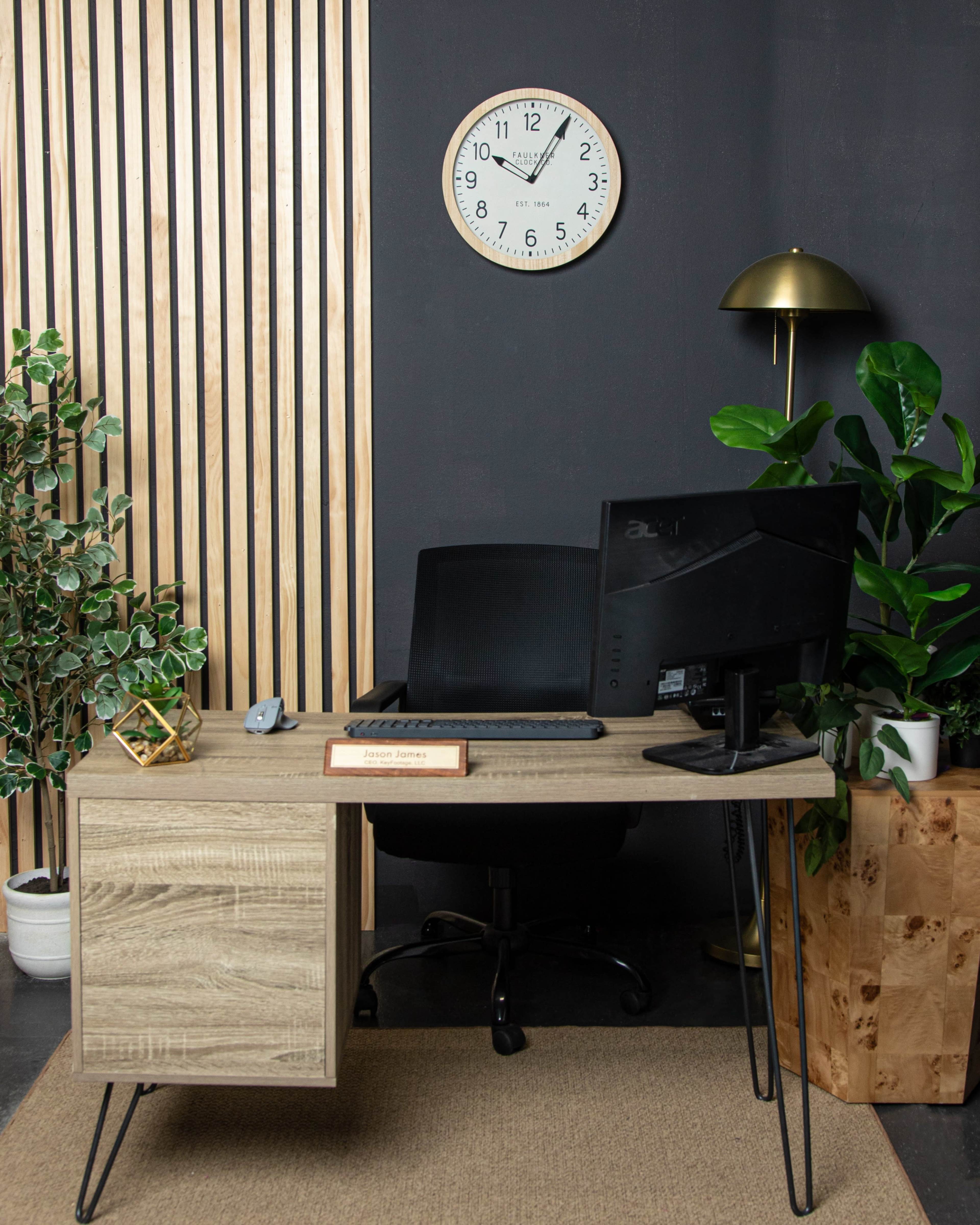 A modern office setup features a wooden desk, an ergonomic chair, a desktop monitor, and decorative plants against a dark wall with wooden paneling and a clock.