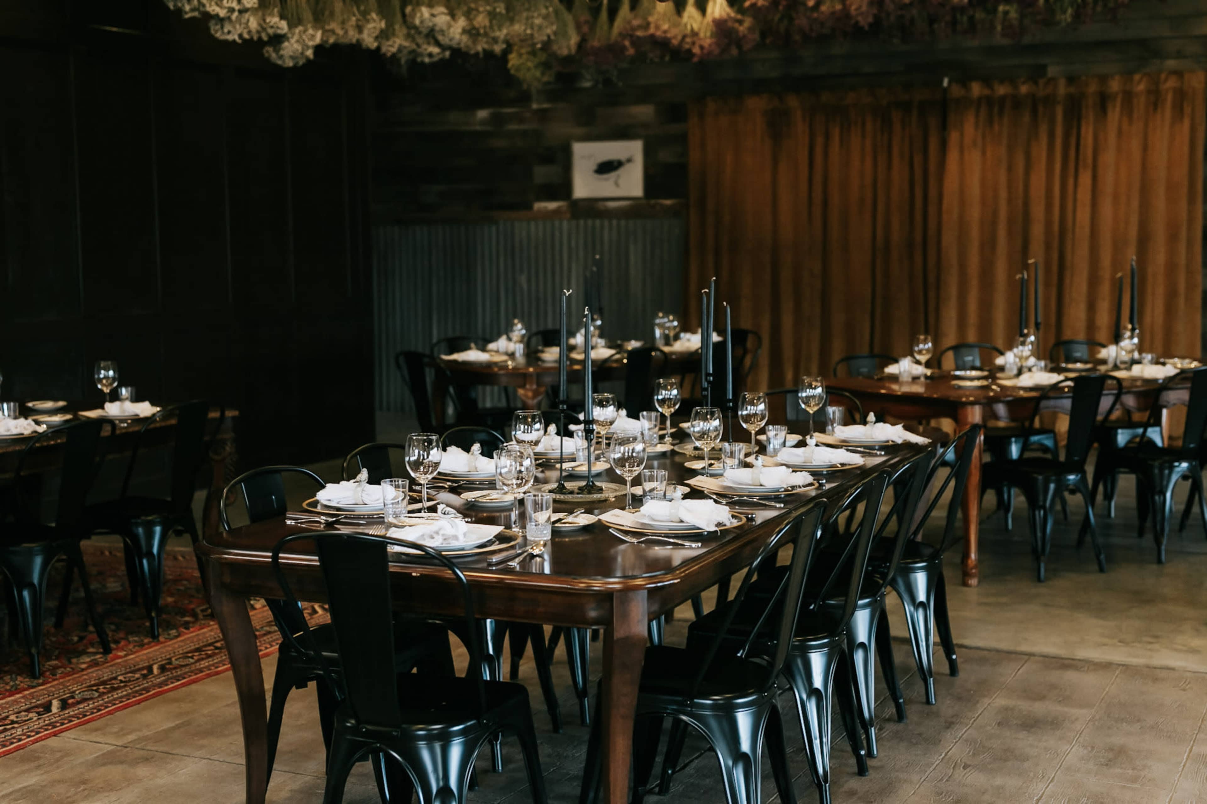 The image shows a dining area with multiple wooden tables set for a formal meal, featuring black chairs and elegant tableware, including glassware and napkins.