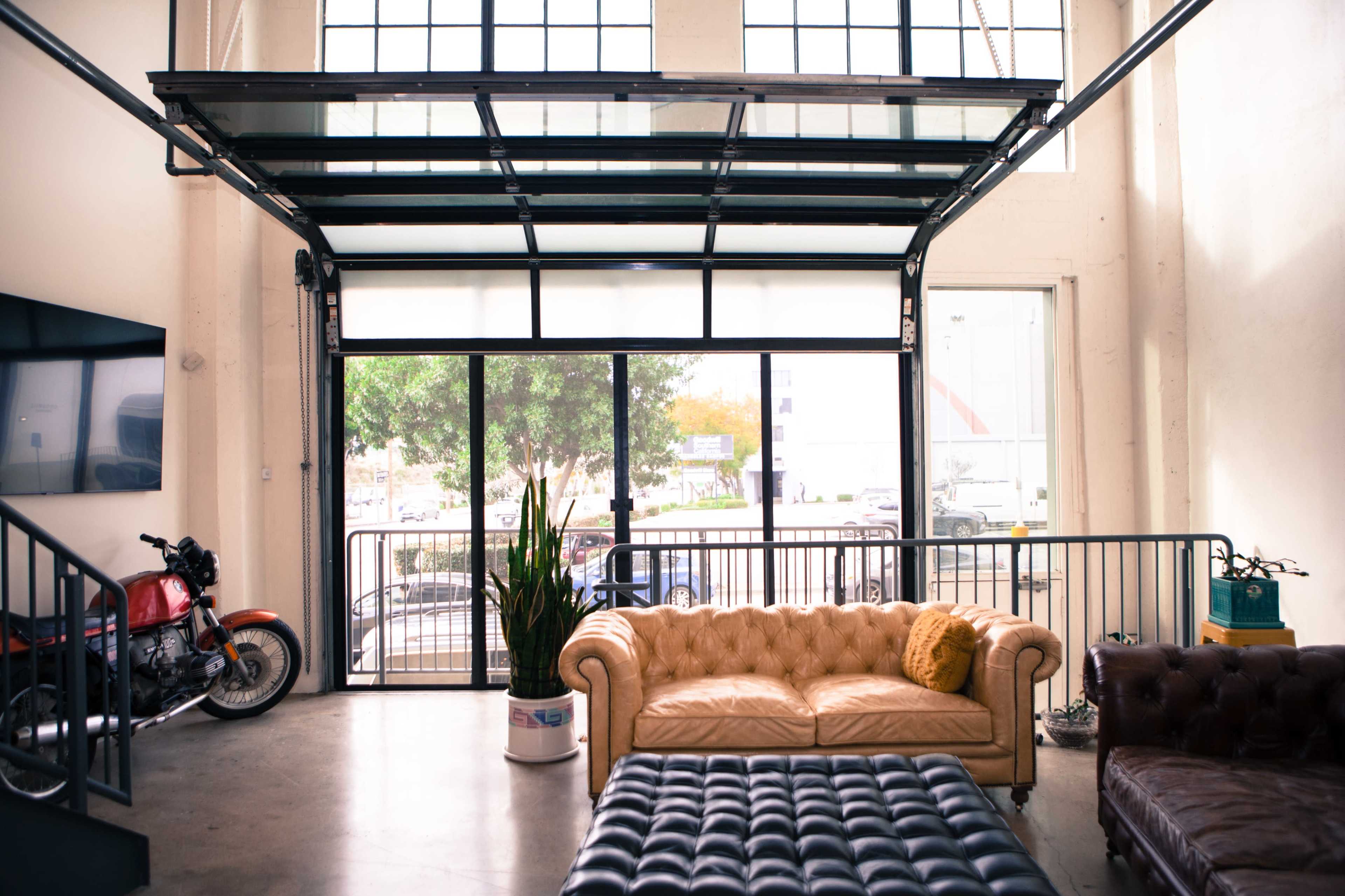 A spacious room with large windows, a beige leather sofa, a dark brown leather couch, and a motorcycle parked near the entrance.