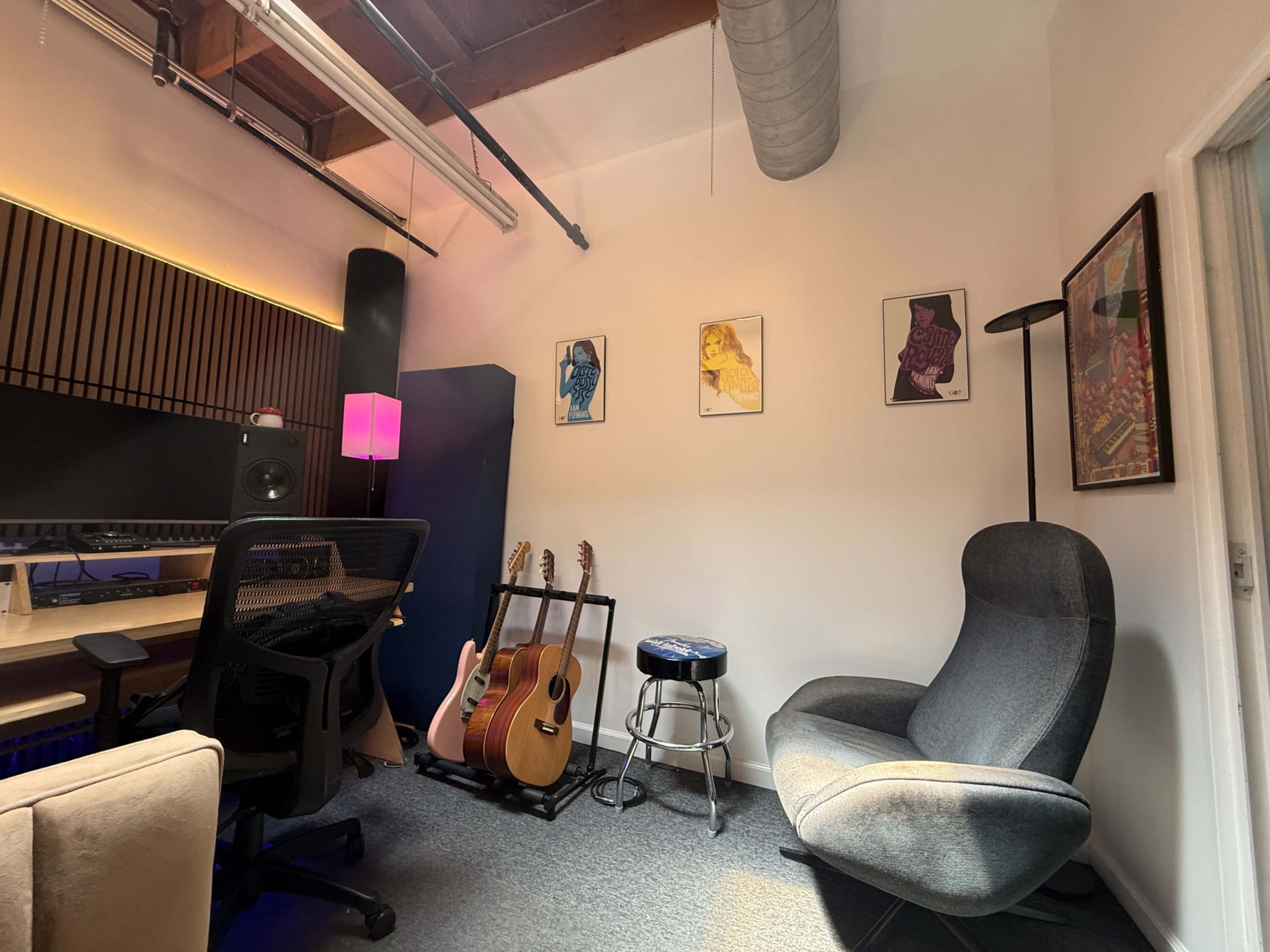 The image shows a music studio featuring a control desk, two guitars on a stand, a stool, and artwork on the walls, with a comfortable chair in the corner.