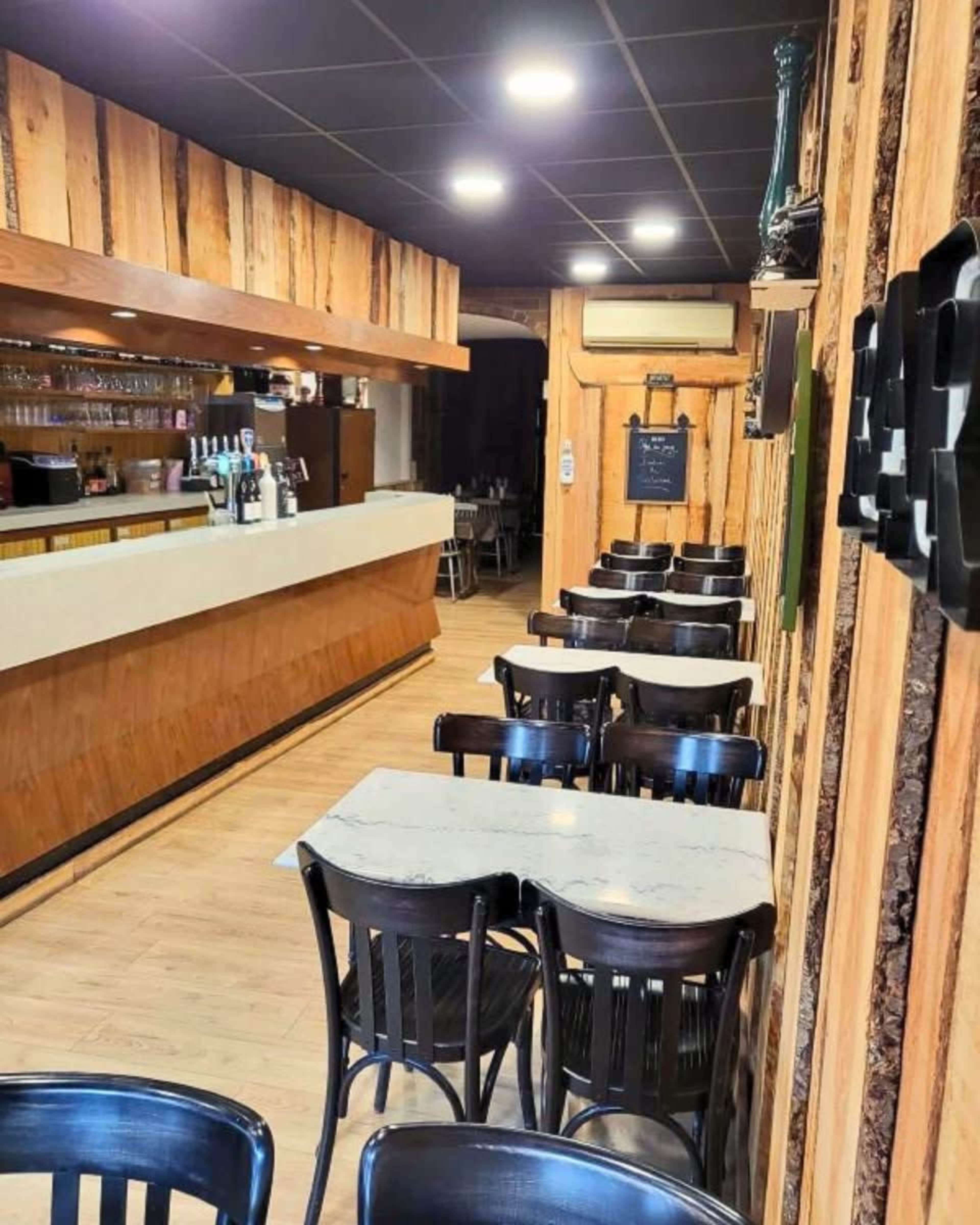 The image shows a modern café with wooden paneling, a bar area, and several black tables and chairs arranged in rows.