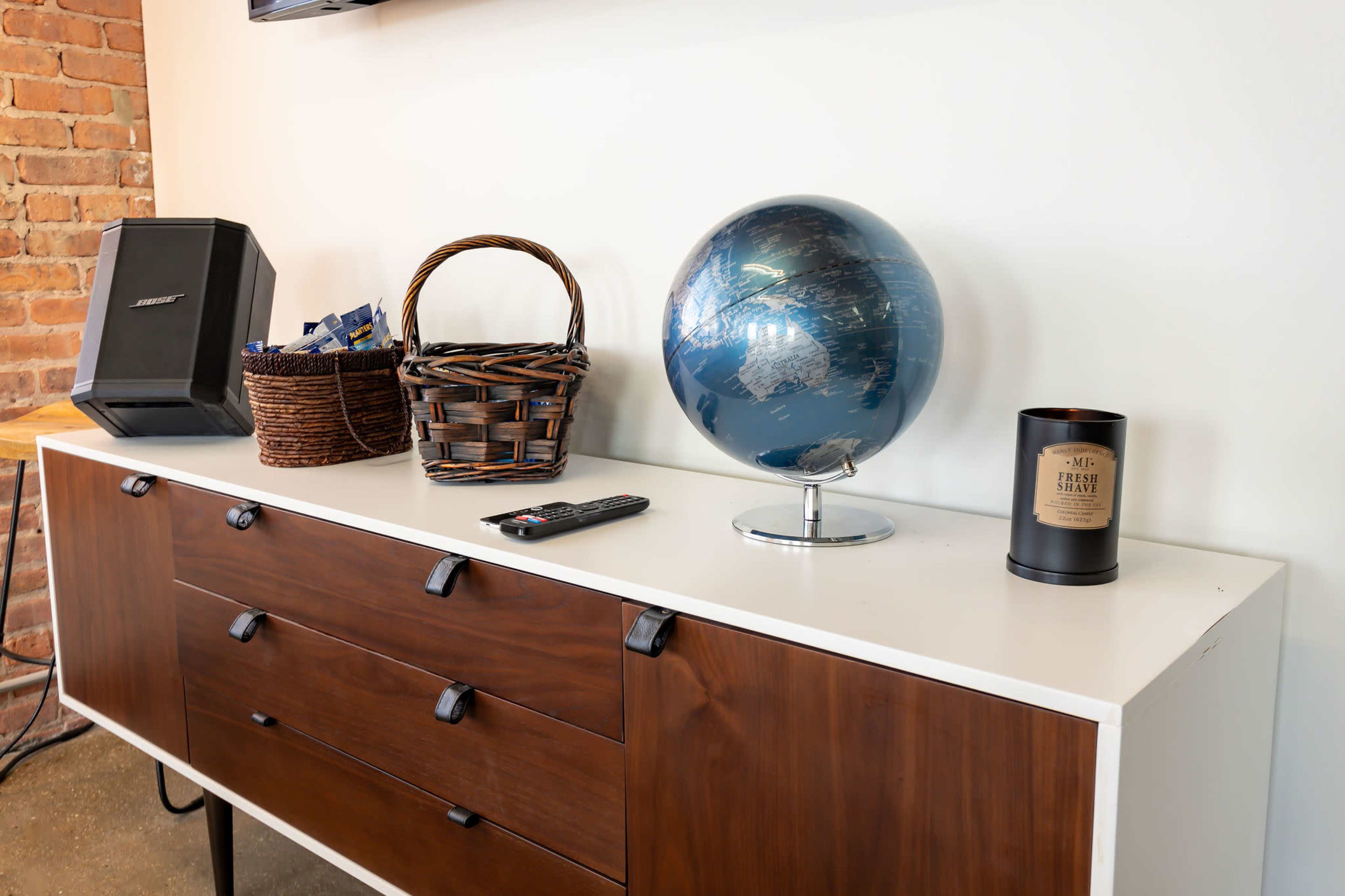 A wooden sideboard displays a globe, a remote control, a decorative candle, a woven basket, and a black speaker against a brick wall.